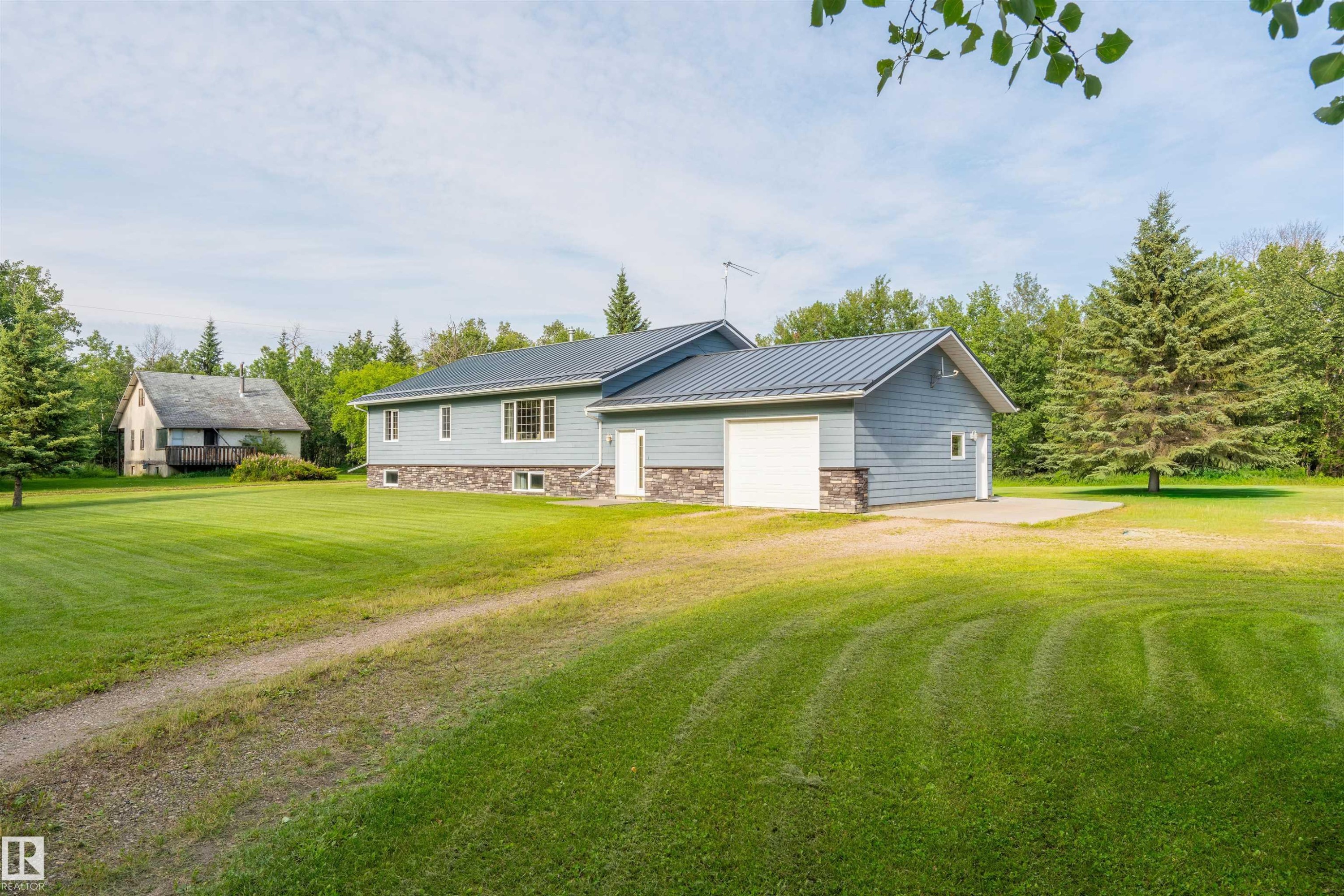 Ranch-style residence featuring light blue siding, a dark metal roof, a garage with a white door, and stone accenting along the foundation - 59125 Range Road 102, Rural St. Paul County, AB - Outdoor