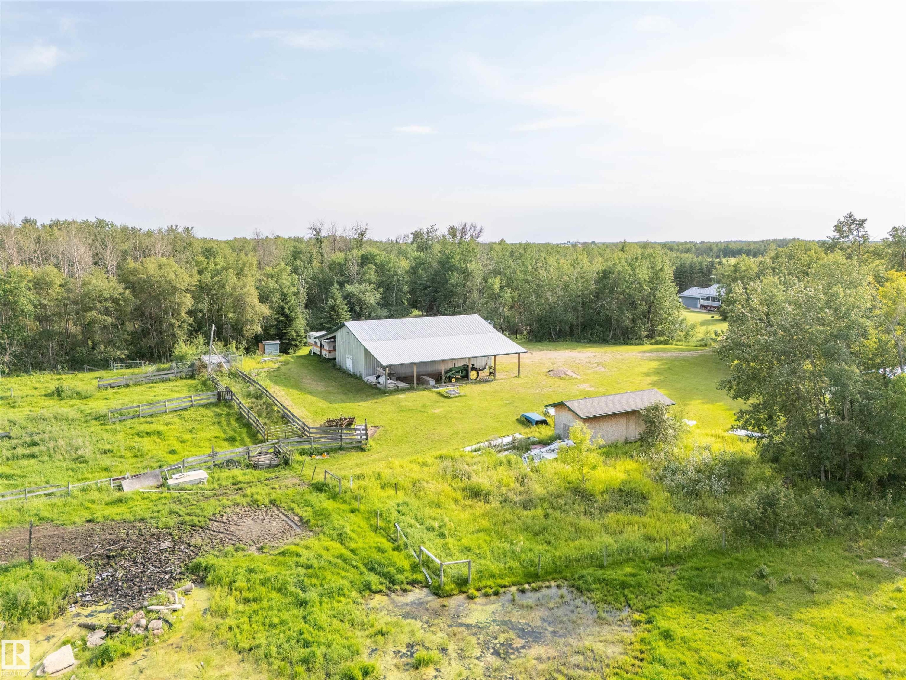 Expansive rural property featuring a large light-colored shop with two attached open-sided structures, a smaller wood-sided outbuilding, fenced pasture areas, and extensive green space surrounded by mature trees - 59125 Range Road 102, Rural St. Paul County, AB - Outdoor With View