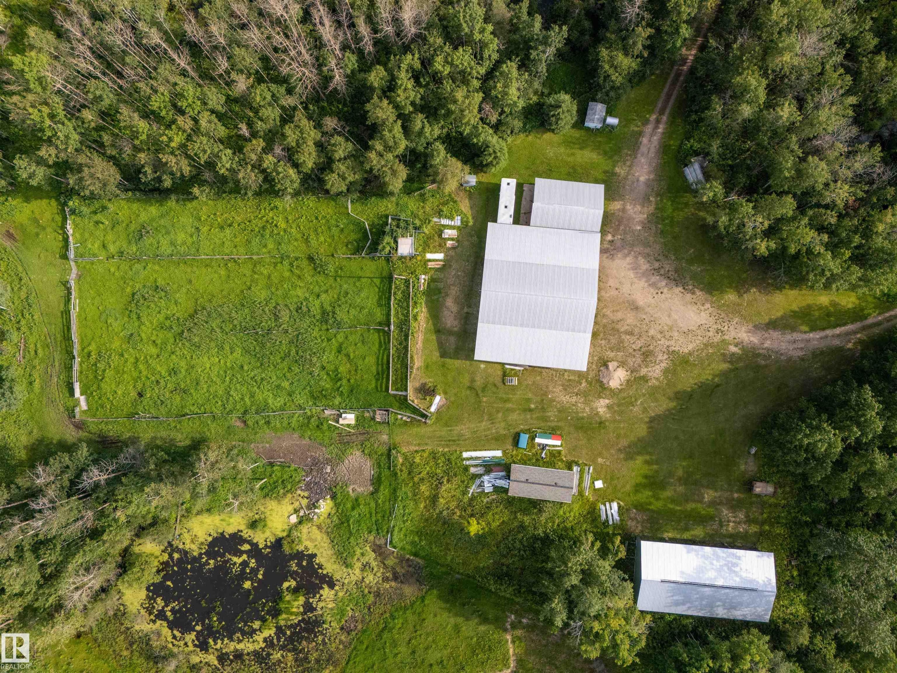 Expansive property featuring a large metal-roofed outbuilding, a smaller metal-roofed structure, a fenced grassy area, a natural pond, and surrounding mature trees - 59125 Range Road 102, Rural St. Paul County, AB - Outdoor