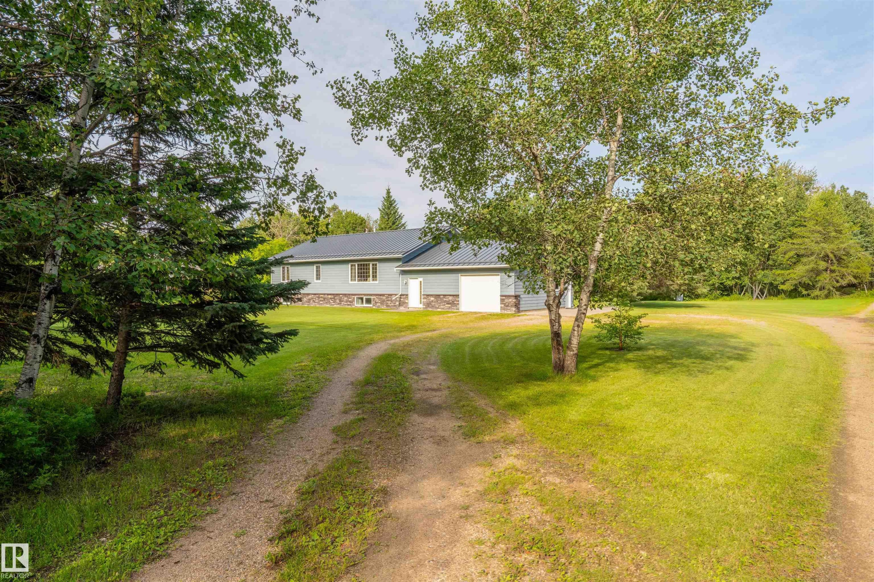 Ranch-style residence featuring light blue siding with a stone foundation, a dark gray metal roof, and an attached garage with white doors - 59125 Range Road 102, Rural St. Paul County, AB - Outdoor