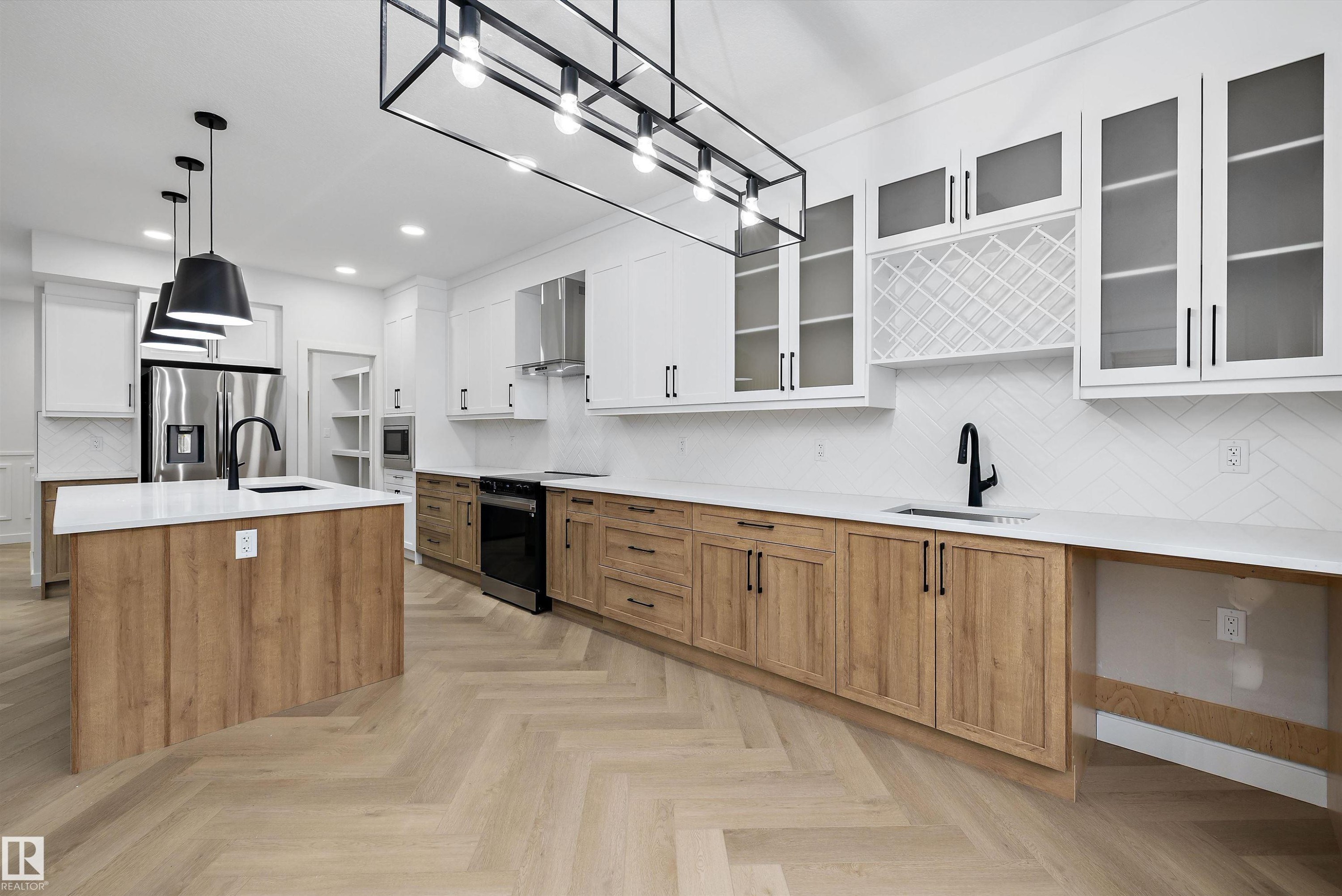 Herringbone wood-finish flooring, an expansive island with a matte black faucet, wood-finish lower cabinetry, white upper cabinetry with glass-front accents, and a distinctive white tile backsplash - 971 Elderberry Landing Nw, Edmonton, AB - Indoor Photo Showing Kitchen