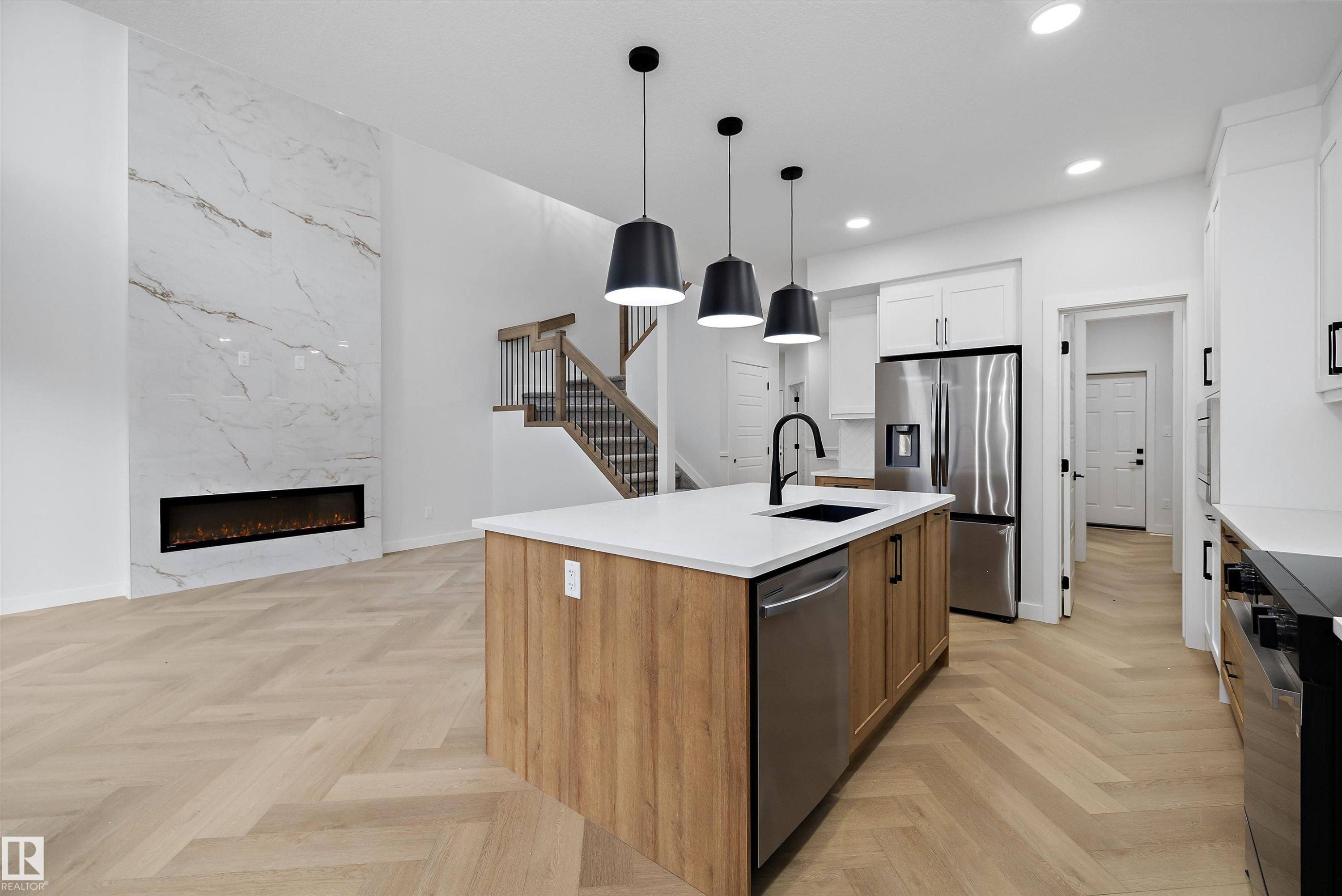 Open-concept living space with herringbone wood-finish flooring, a full-height fireplace with a linear insert, and a kitchen island featuring a white countertop and pendant lighting - 971 Elderberry Landing Nw, Edmonton, AB - Indoor Photo Showing Kitchen With Upgraded Kitchen