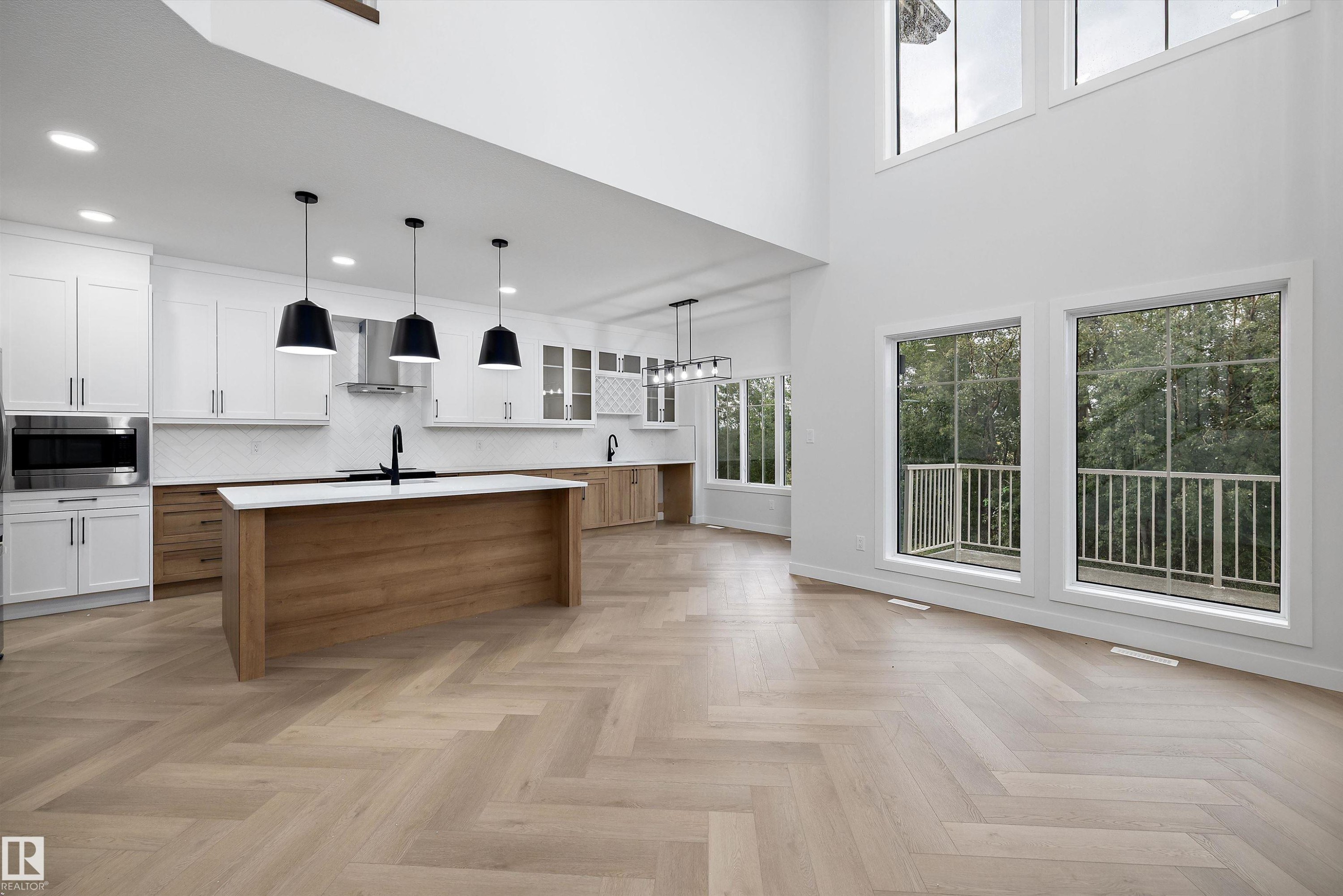 Herringbone wood-finish flooring, two-story living space with multiple windows, kitchen with white upper cabinetry and wood-finish lower cabinetry, kitchen island with built-in sink, and pendant lighting - 971 Elderberry Landing Nw, Edmonton, AB - Indoor Photo Showing Other Room