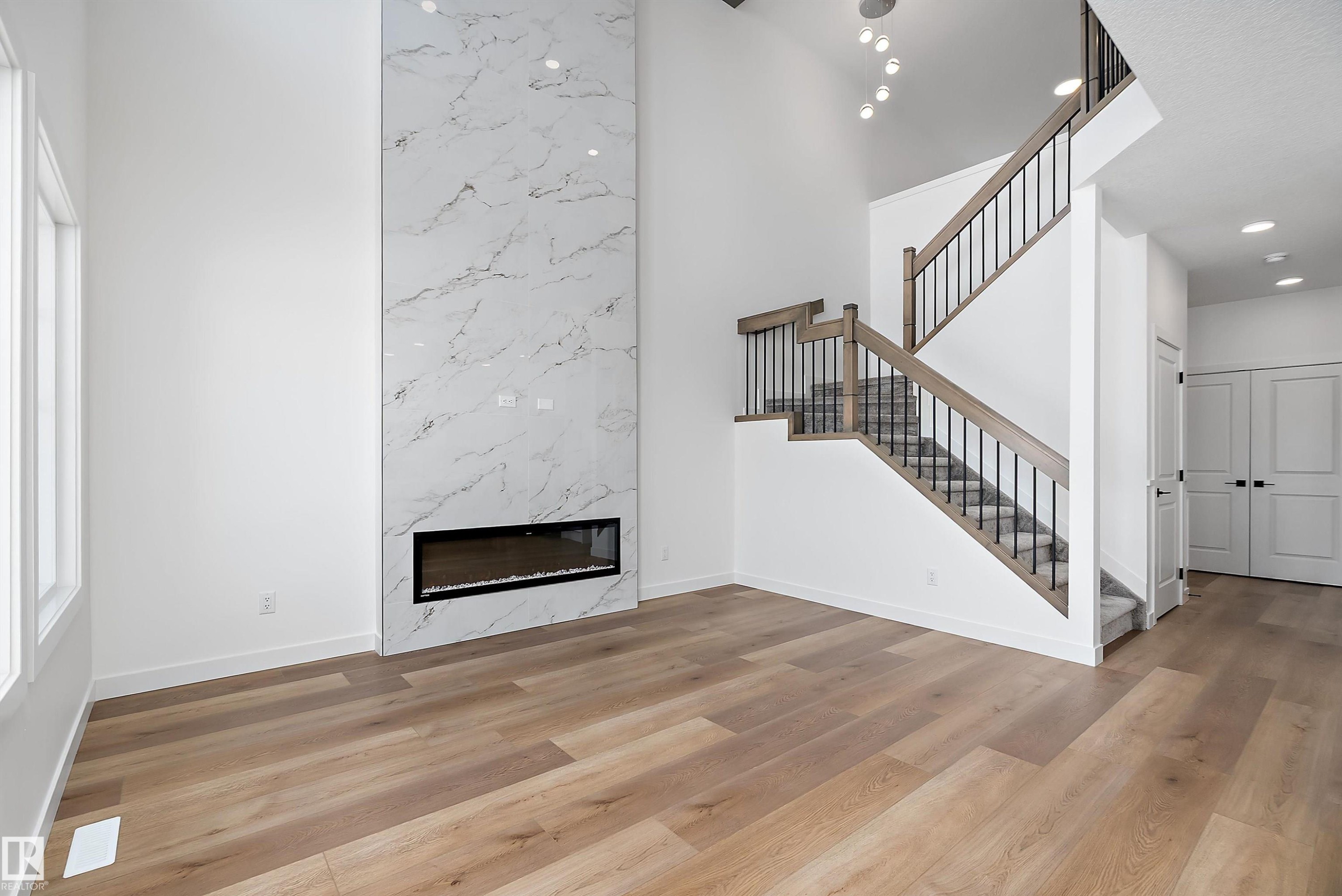Two-story interior with wood-finish flooring, a linear electric fireplace with a marble-style surround, and a staircase with wood handrails and black metal balusters - 1763 Crossbill Loop Nw, Edmonton, AB - Indoor With Fireplace