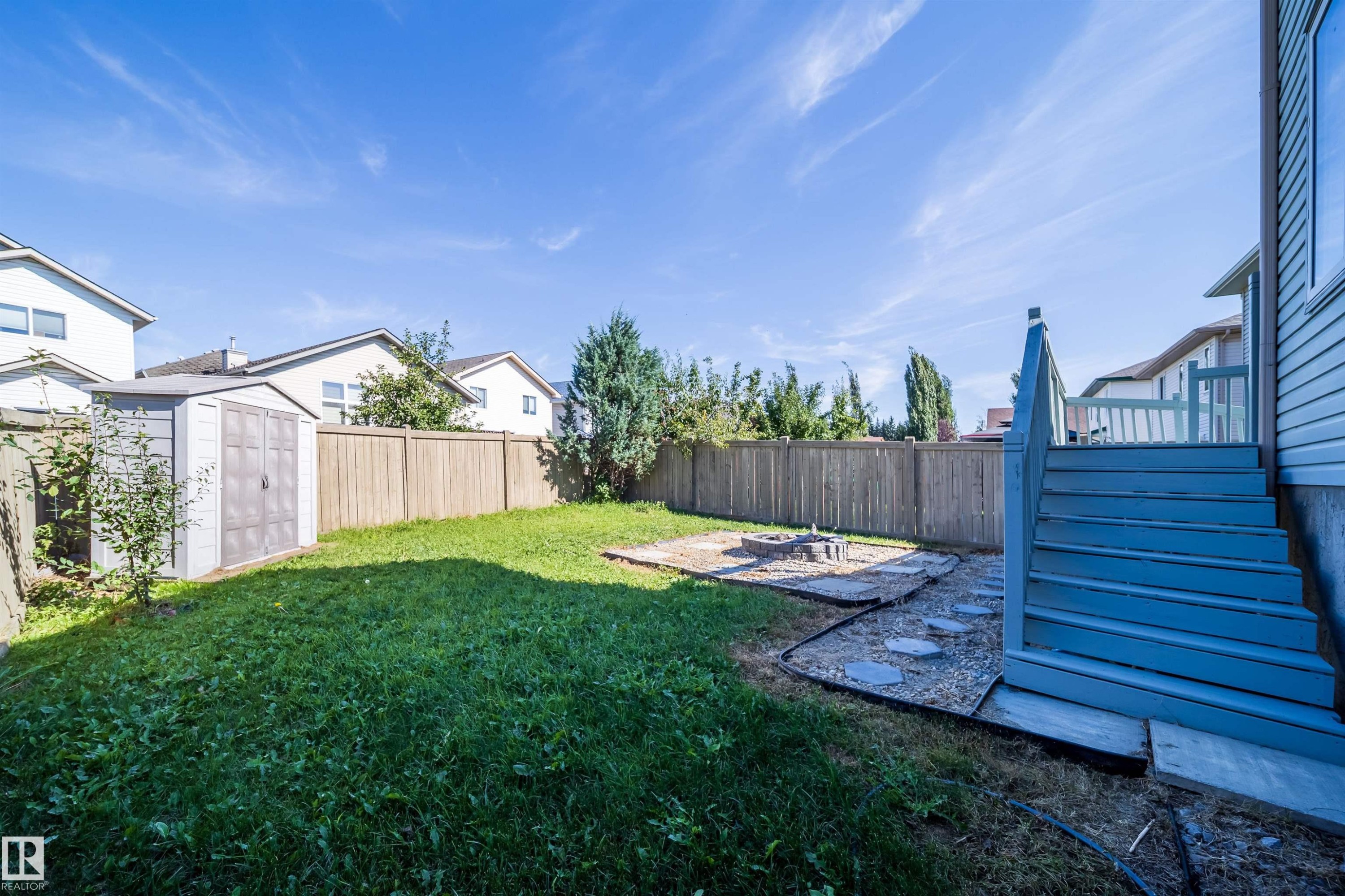 Expansive backyard featuring a wood fence, storage shed, raised deck with stairs, and a stone-paved area - 7848 7 Avenue Sw, Edmonton, AB