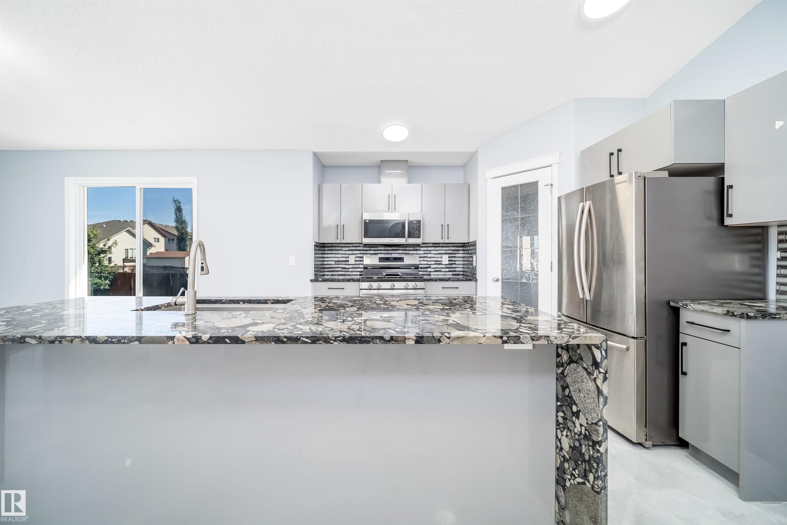 Kitchen featuring an expansive island with a polished countertop, stainless steel appliances, modern cabinetry, a tile backsplash, and a sliding glass door - 7848 7 Avenue Sw, Edmonton, AB