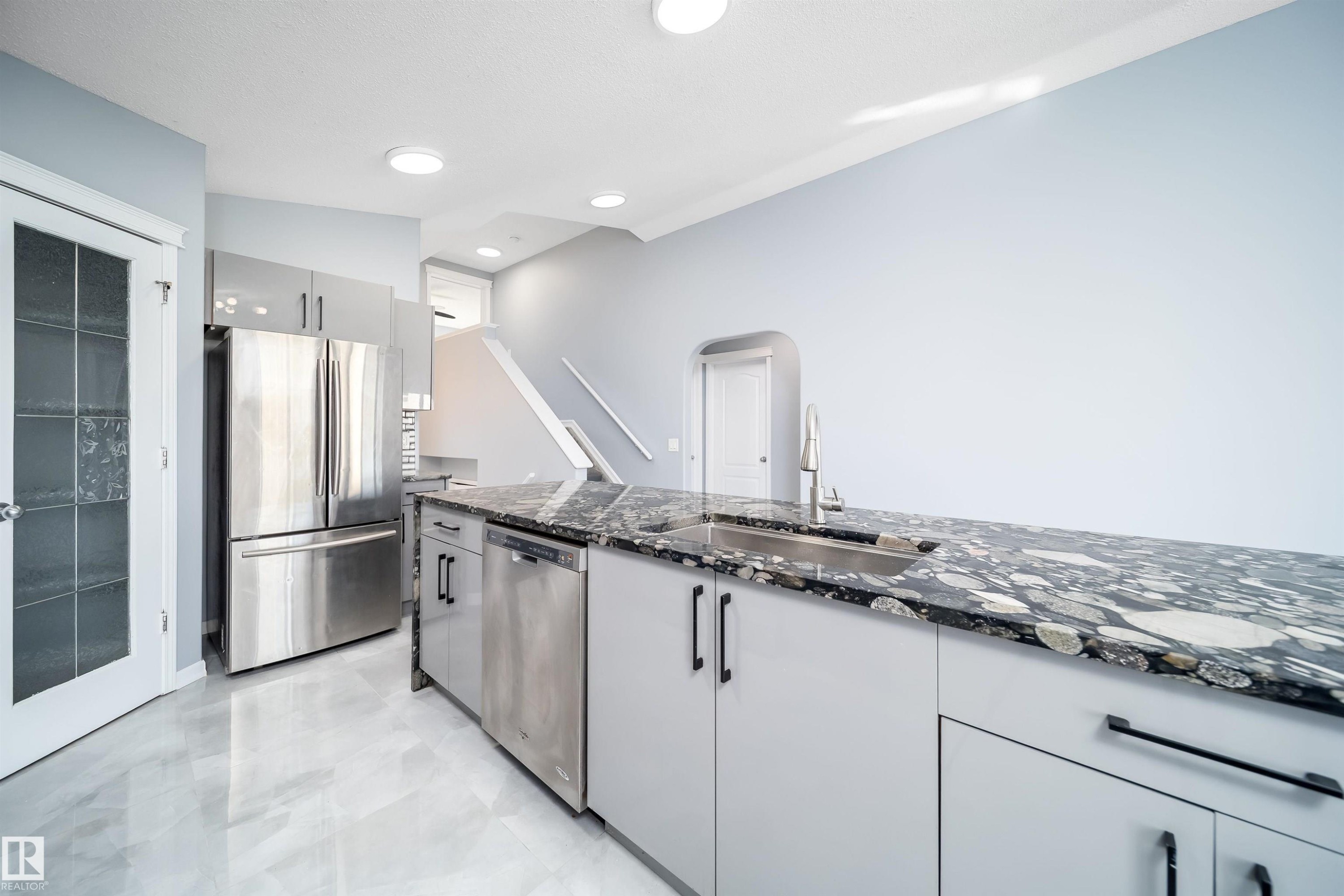Kitchen featuring a stainless steel refrigerator, stainless steel dishwasher, light gray cabinetry, dark patterned countertop, and an undermount sink with a gooseneck faucet - 7848 7 Avenue Sw, Edmonton, AB