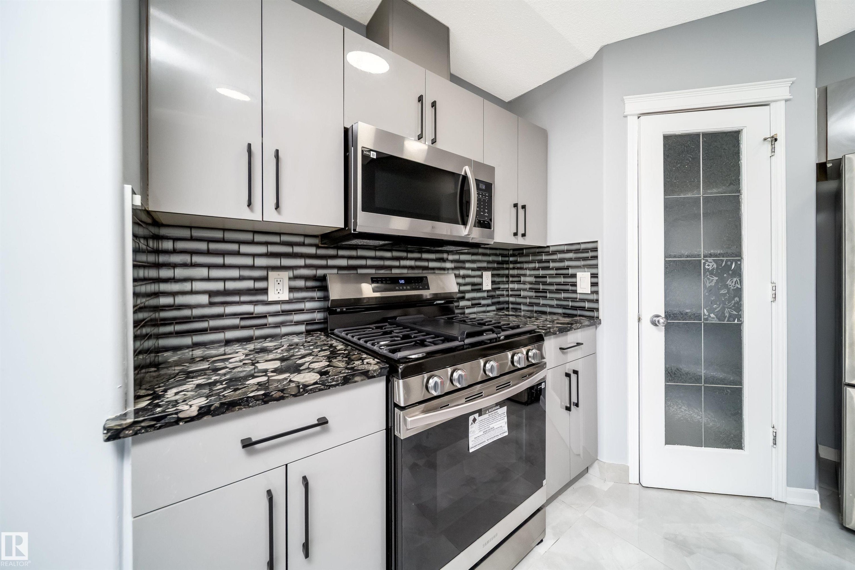 Kitchen featuring light grey cabinetry with dark hardware, stainless steel appliances, a tile backsplash, and patterned countertops - 7848 7 Avenue Sw, Edmonton, AB