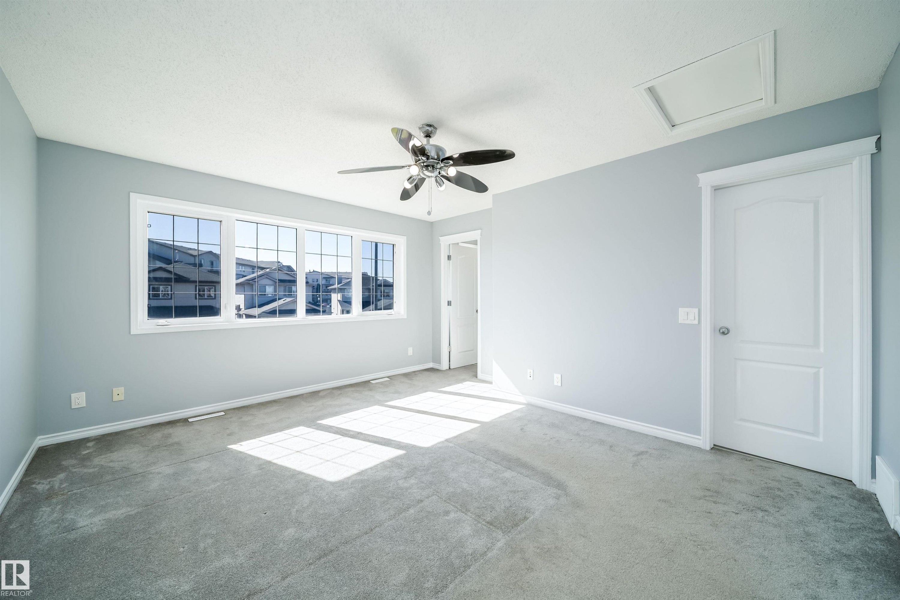 Spacious room featuring light gray carpeting, a multi-panel window with grid accents, a ceiling fan with light fixture, and a white paneled door - 7848 7 Avenue Sw, Edmonton, AB