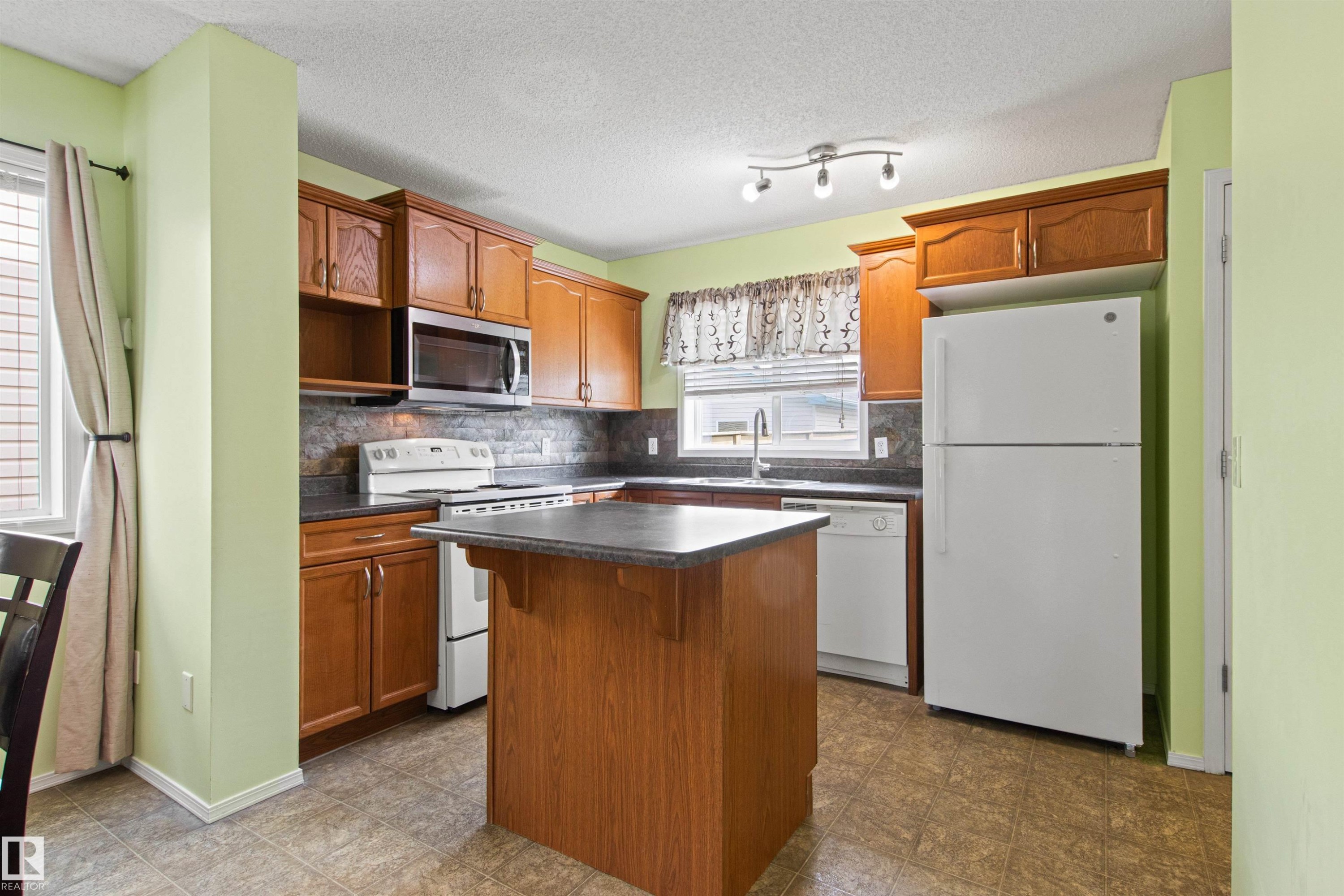 Kitchen featuring wood-finish cabinetry, a central island with a dark countertop, tile flooring, and white appliances including a refrigerator, range, and dishwasher - 9118 Scott Crescent Nw, Edmonton, AB - Indoor Photo Showing Kitchen