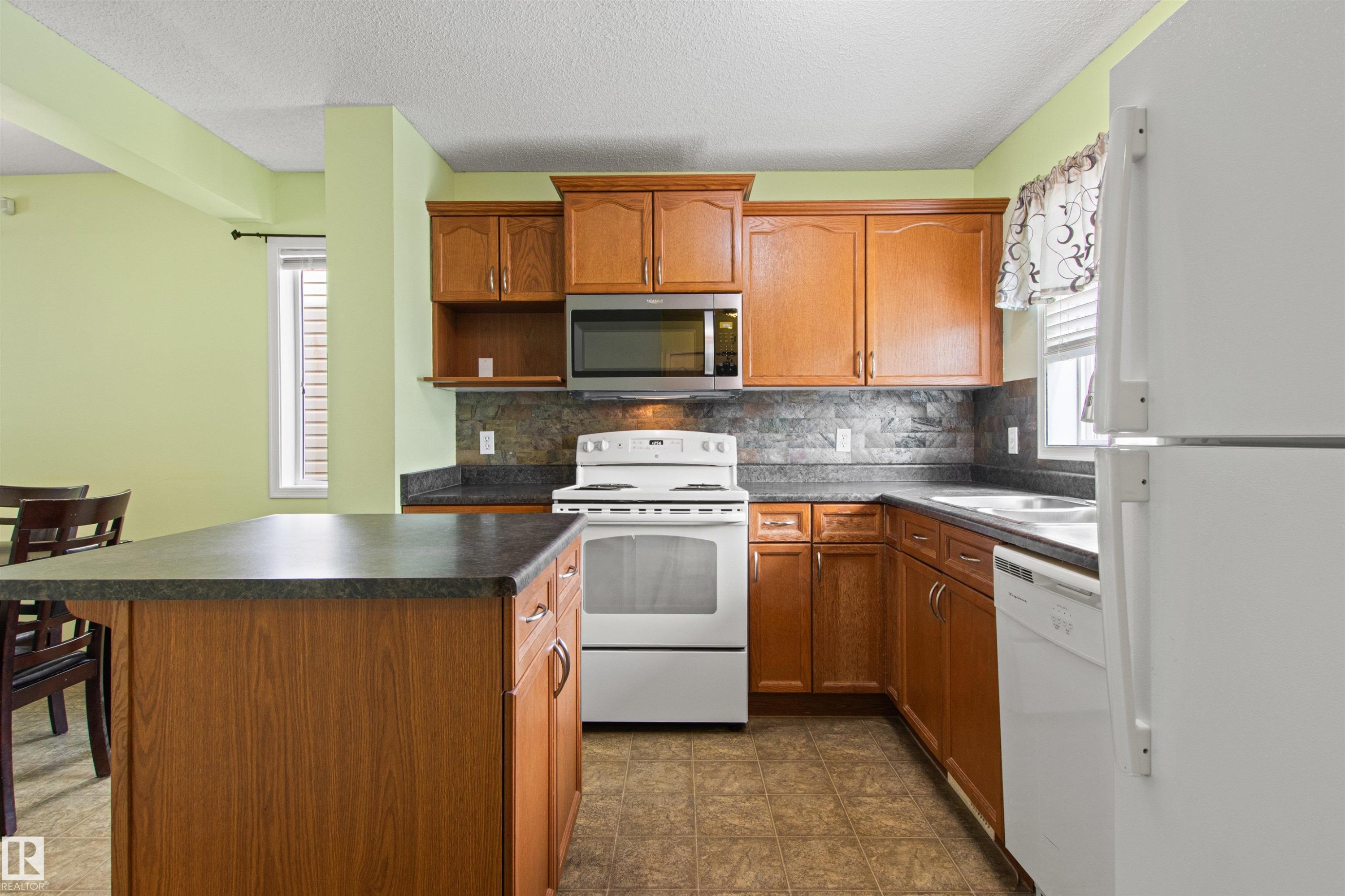 Kitchen featuring wood-finish cabinetry, a central island with a dark countertop, white appliances, and a tiled backsplash - 9118 Scott Crescent Nw, Edmonton, AB - Indoor Photo Showing Kitchen
