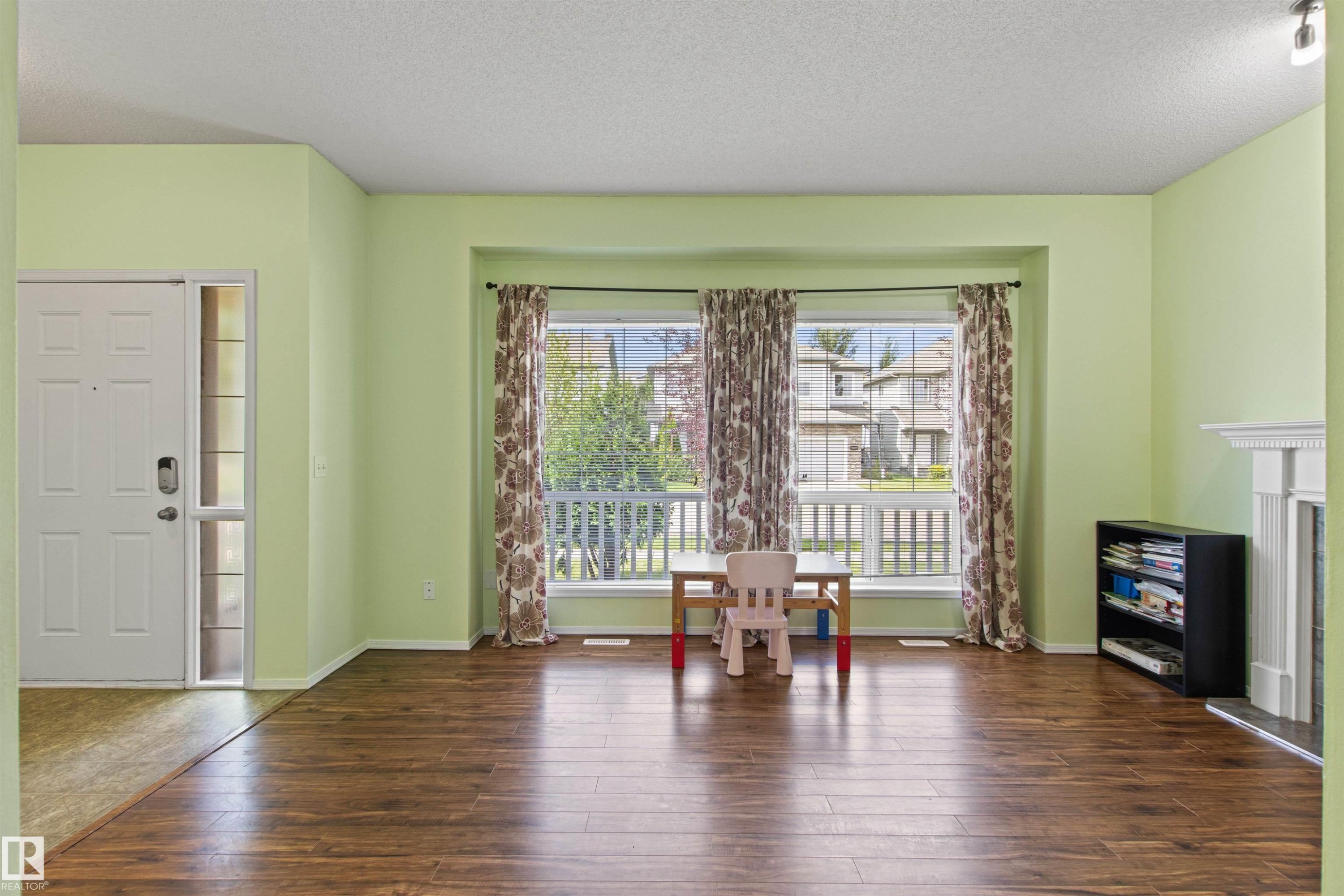 Wood-finish flooring, white entry door with frosted glass sidelight, bay window with white trim, and a decorative white fireplace surround - 9118 Scott Crescent Nw, Edmonton, AB - Indoor Photo Showing Other Room