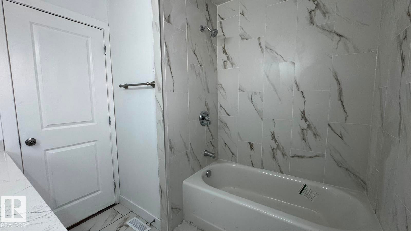 Bathtub with a shower, white and gray veined tile surround, chrome fixtures, and a white paneled door - 41 Lancaster Terrace Nw, Edmonton, AB - Indoor Photo Showing Bathroom