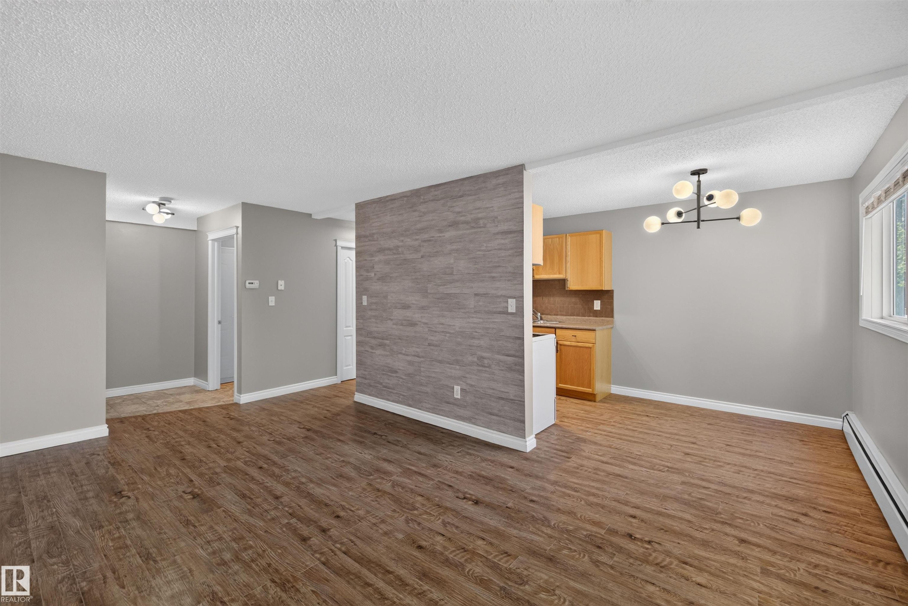 Wood-finish flooring, accent wall with textured finish, modern multi-bulb chandelier, kitchen area with wood cabinetry, and a window with white trim - #4 4616 47 St, Leduc, AB - Indoor