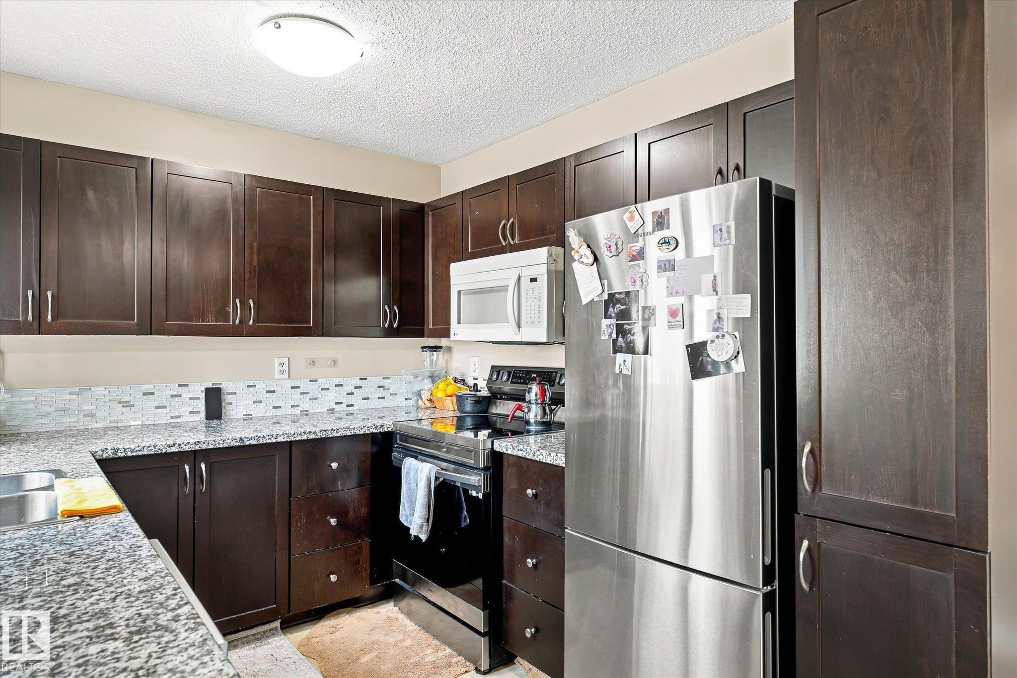 Dark wood-finish cabinetry, stainless steel appliances, granite-style countertops, and a mosaic tile backsplash - 6471 180 Street Nw, Edmonton, AB - Indoor Photo Showing Kitchen With Stainless Steel Kitchen