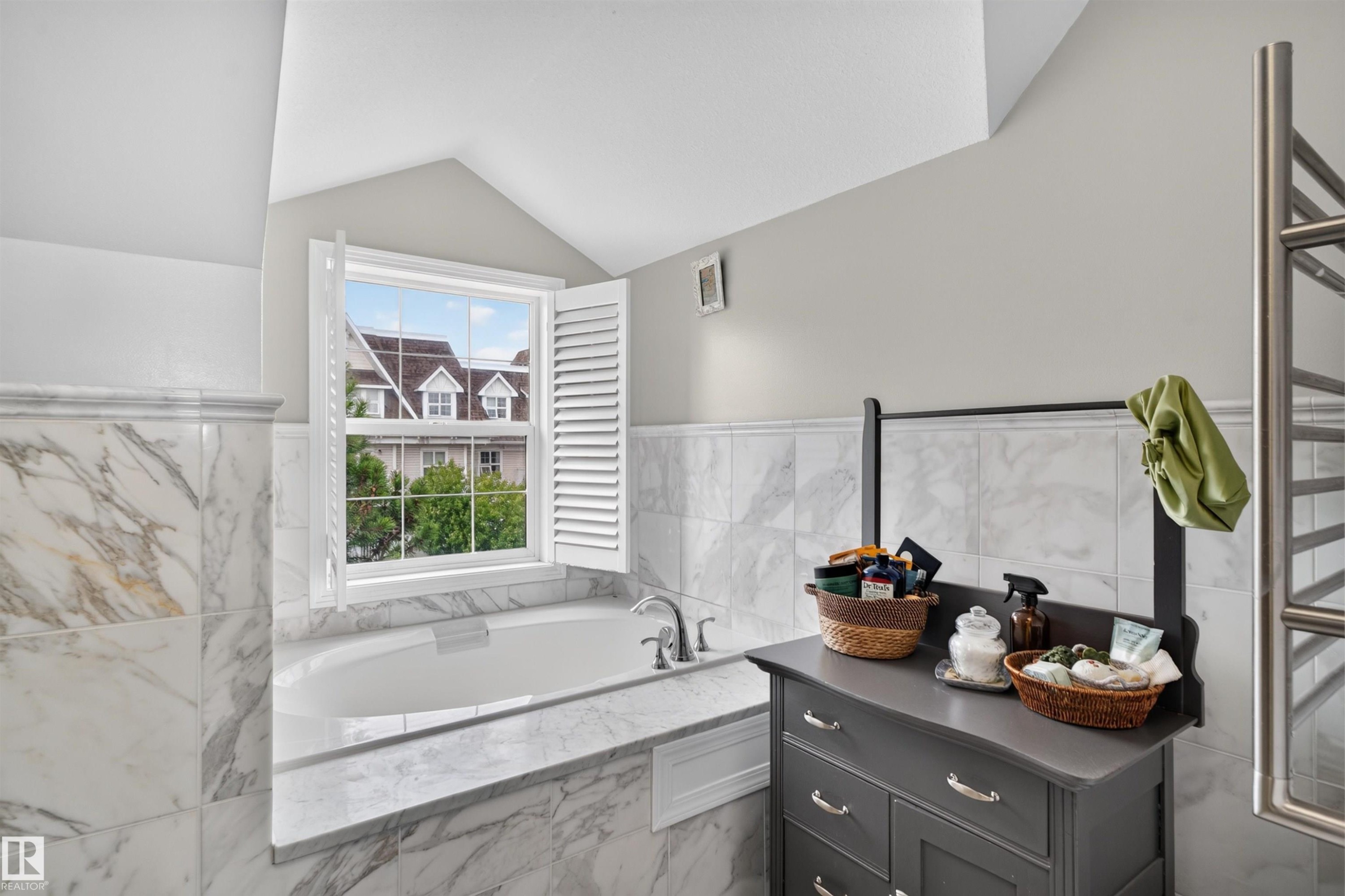 Built-in soaking tub, white plantation shutters, white window frame, marble-finish tile surround, and a wall-mounted towel warmer - 4471 Mccrae Avenue Nw, Edmonton, AB
