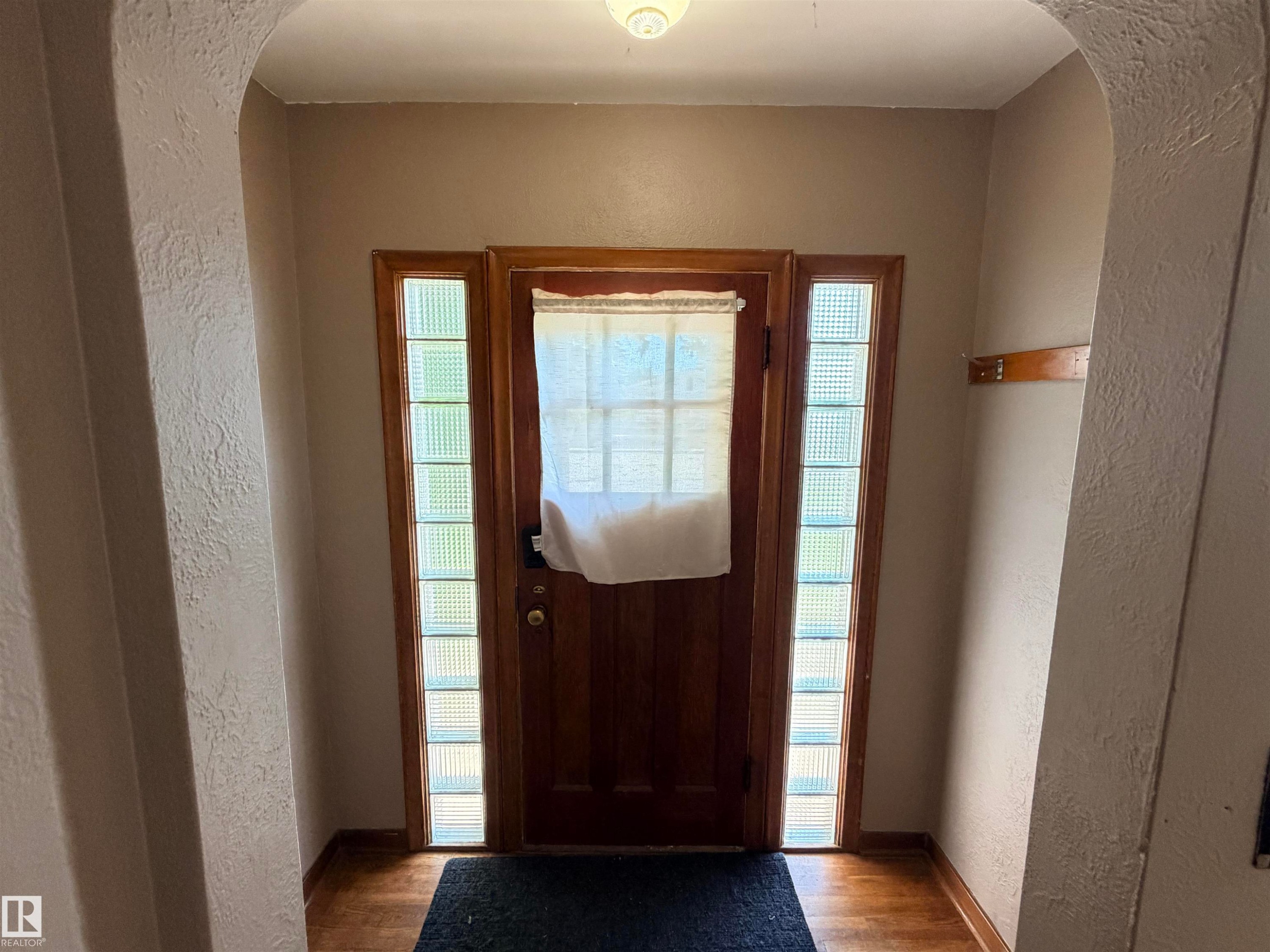 Wood-finish flooring with a dark area rug, featuring a solid wood door flanked by glass block sidelights - 10416 100 Avenue, Westlock, AB - Indoor Photo Showing Other Room