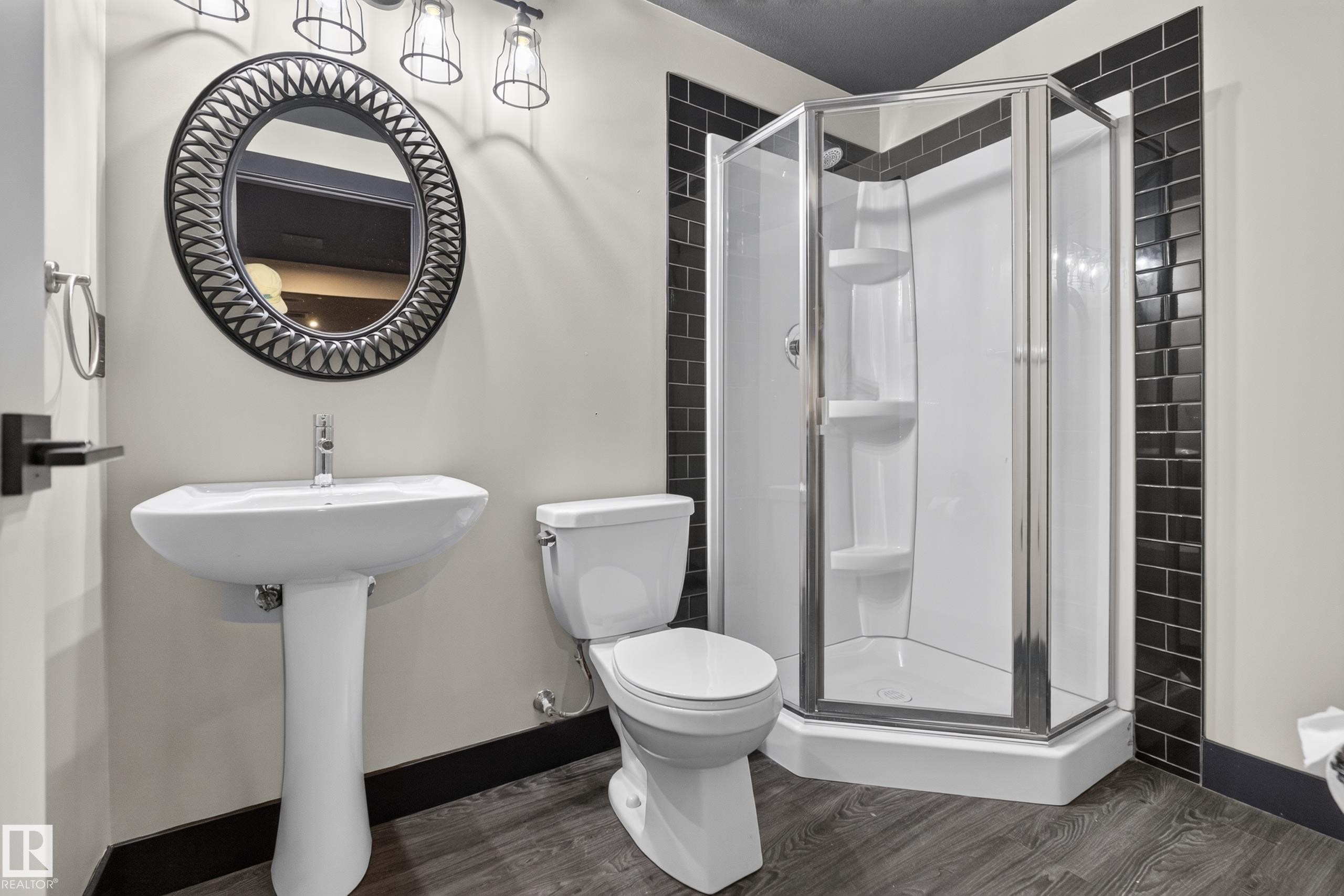 Pedestal sink with chrome faucet, round decorative mirror, corner shower enclosure with built-in shelving, wood-finish flooring, and subway tile accents - 1399 Graydon Hill Way Sw, Edmonton, AB