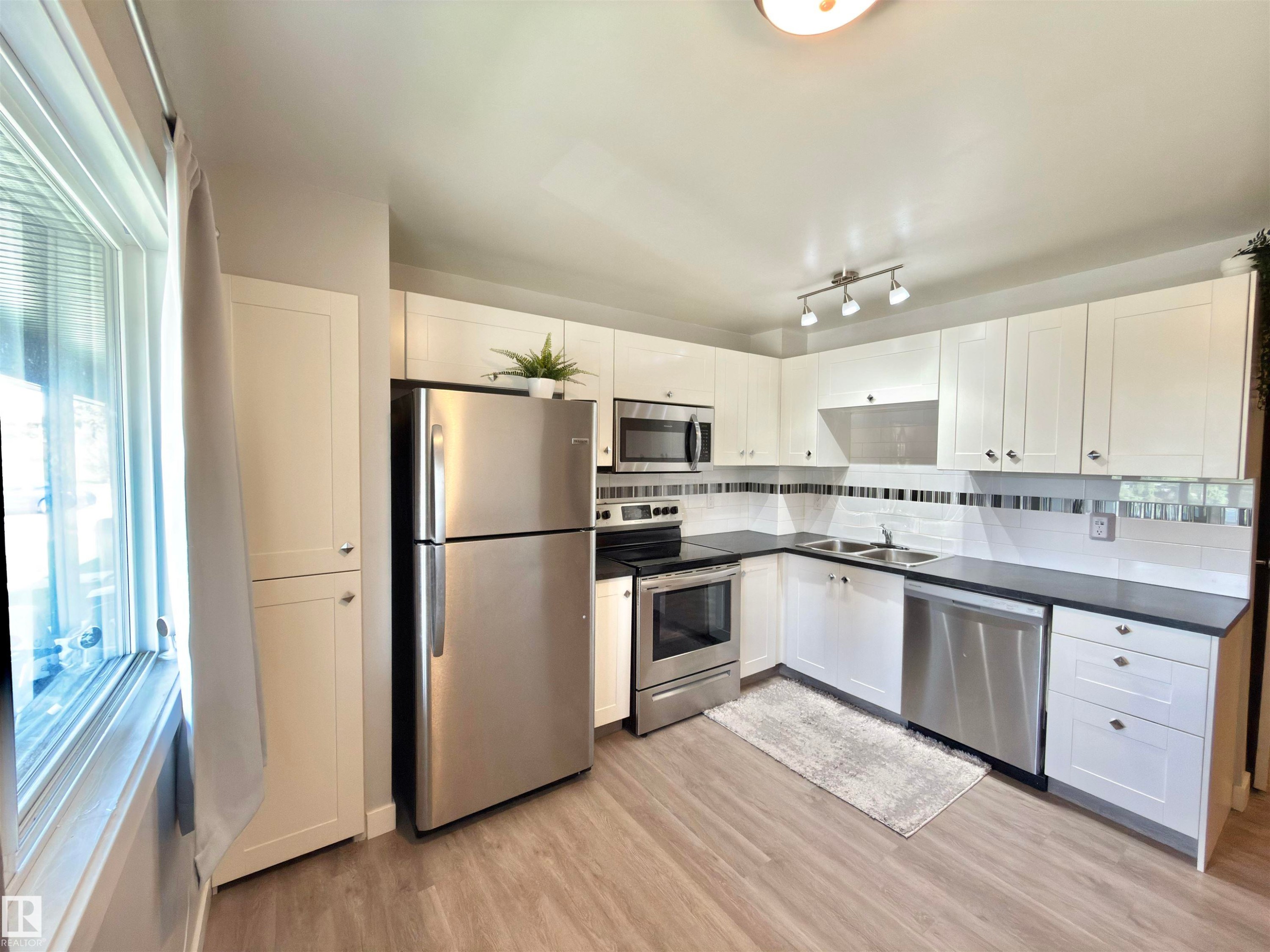 Kitchen featuring white cabinetry, stainless steel appliances, dark countertops, white tile backsplash with metallic accents, and wood-finish flooring - #13 18010 98 Av Nw Nw, Edmonton, AB - Indoor Photo Showing Kitchen With Stainless Steel Kitchen With Double Sink