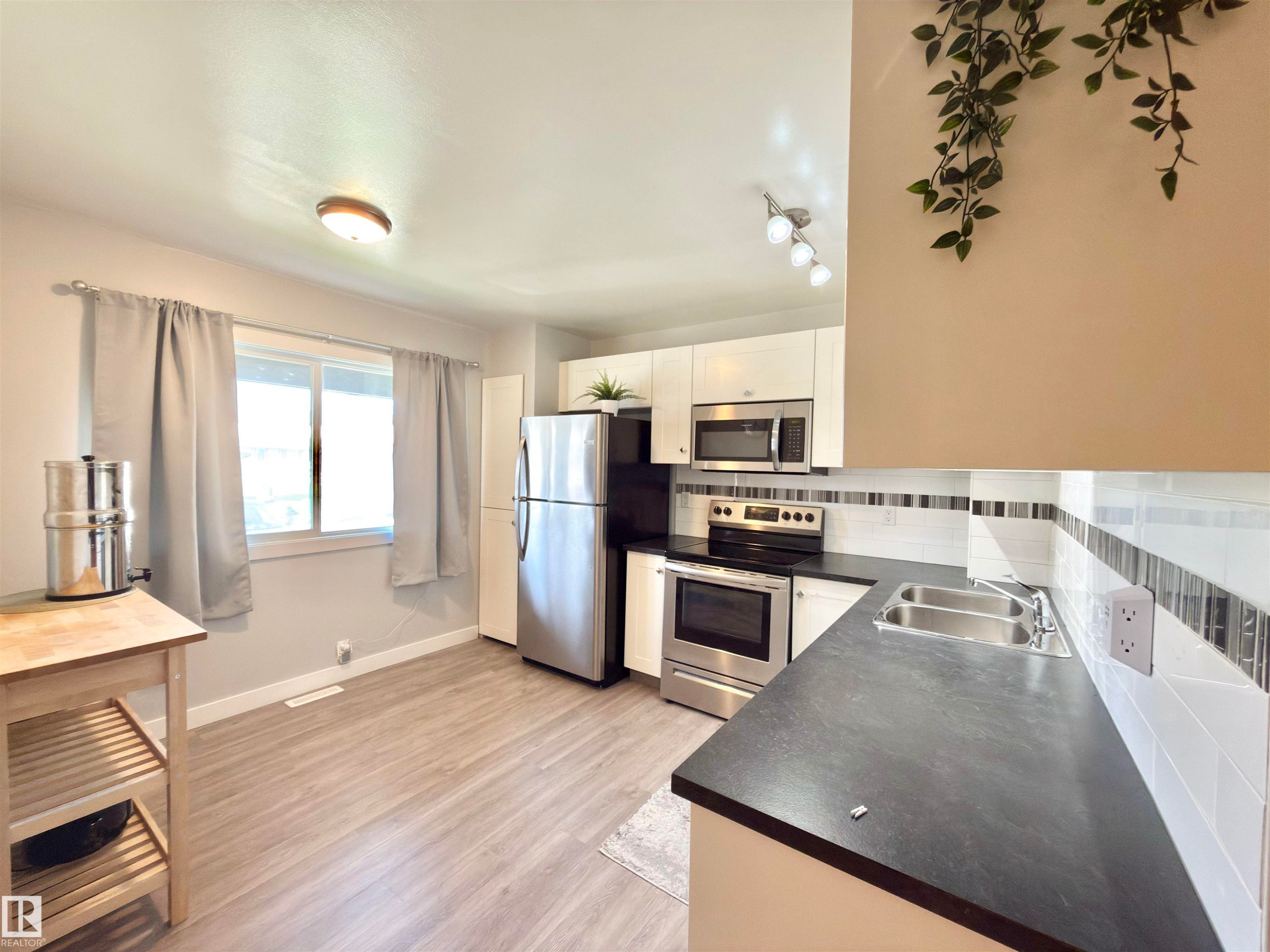 Kitchen featuring stainless steel appliances, white cabinetry, dark countertops, a white subway tile backsplash with accent tiling, and wood-finish flooring - #13 18010 98 Av Nw Nw, Edmonton, AB - Indoor Photo Showing Kitchen With Stainless Steel Kitchen With Double Sink