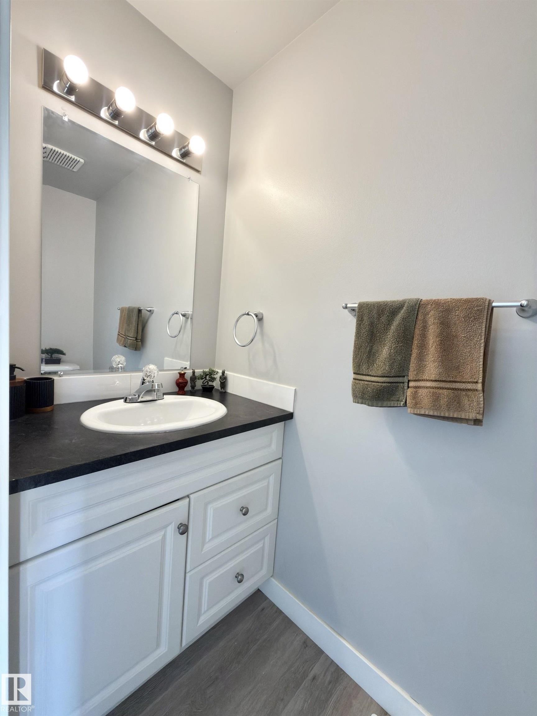 White vanity with a dark countertop and integrated sink, chrome fixtures, and wood-finish flooring - #13 18010 98 Av Nw Nw, Edmonton, AB - Indoor Photo Showing Bathroom
