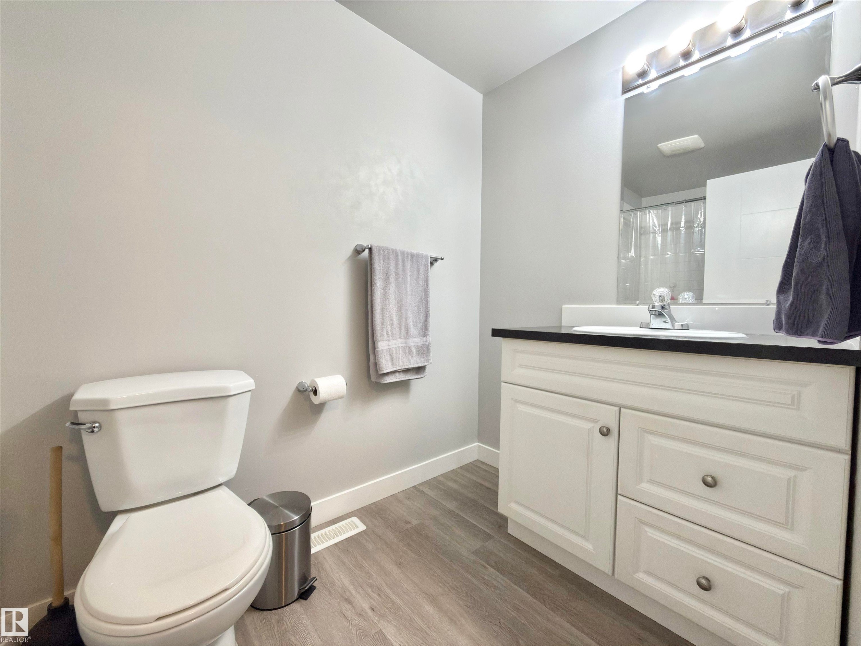 Bathroom featuring a white vanity with cabinet storage, black countertop, mirrored medicine cabinet with integrated lighting, and wood-finish flooring - #13 18010 98 Av Nw Nw, Edmonton, AB - Indoor Photo Showing Bathroom