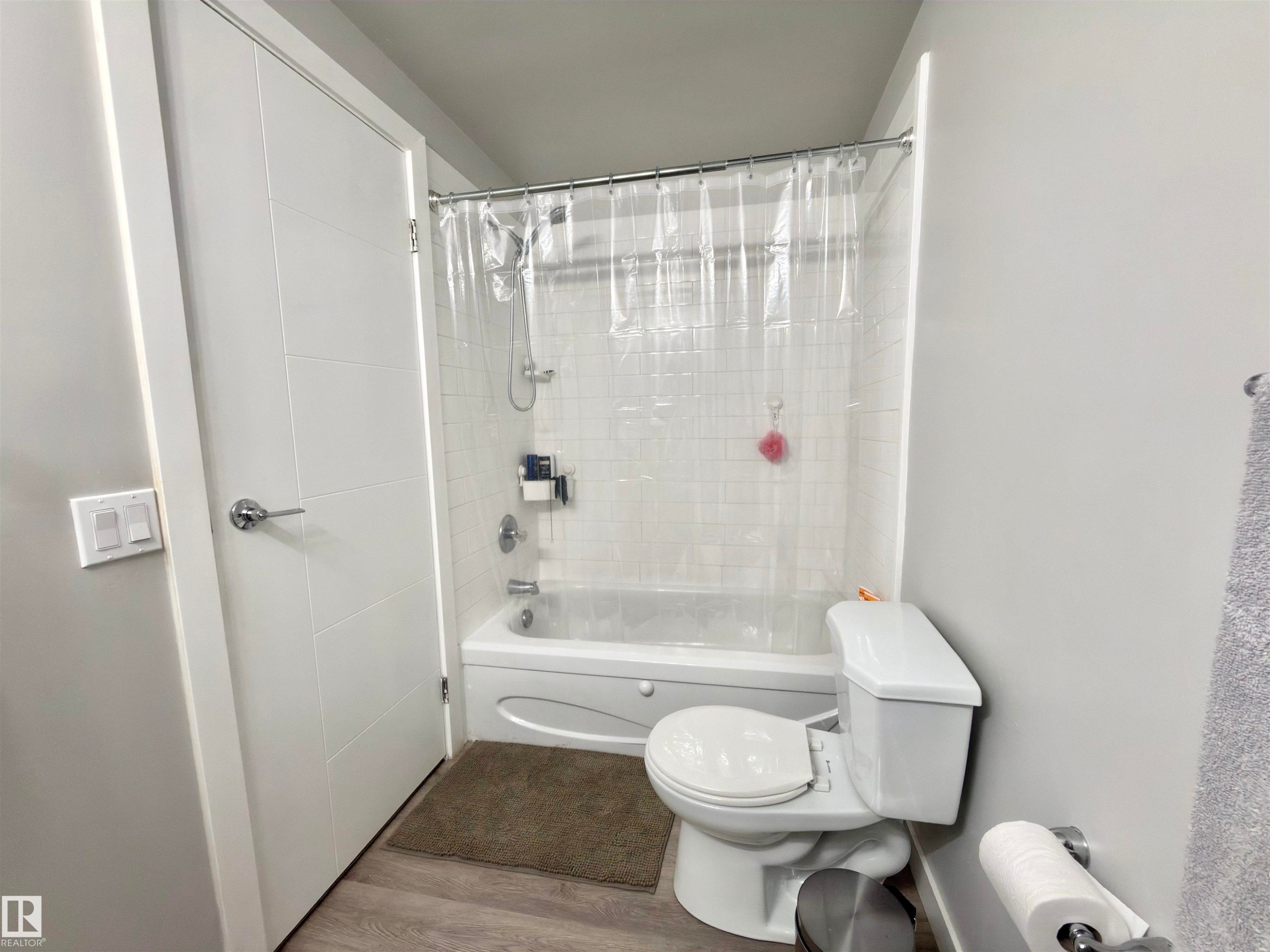 Bathtub with subway tile surround, white toilet, chrome fixtures, and wood-finish flooring - #13 18010 98 Av Nw Nw, Edmonton, AB - Indoor Photo Showing Bathroom