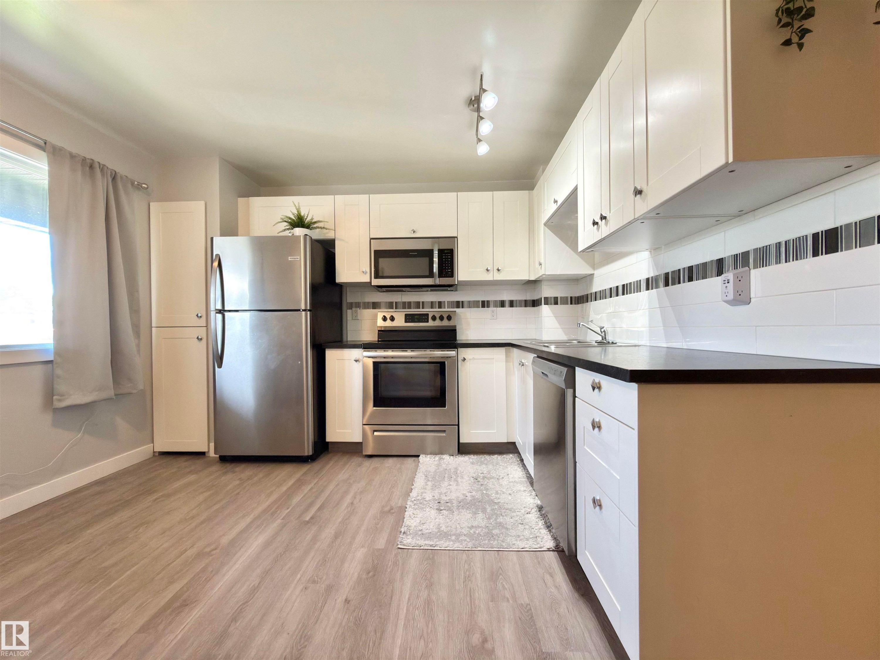 Kitchen featuring white cabinetry, dark countertops, a white subway tile backsplash with accent stripe, stainless steel appliances, and wood-finish flooring - #13 18010 98 Av Nw Nw, Edmonton, AB - Indoor Photo Showing Kitchen With Stainless Steel Kitchen