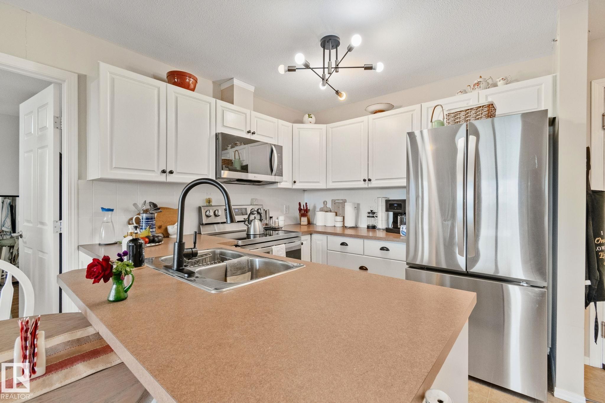Kitchen featuring white cabinetry, stainless steel appliances, a double basin sink with a gooseneck faucet, and a contemporary chandelier - #407 4309 33 St, Stony Plain, AB - Indoor Photo Showing Kitchen With Double Sink With Upgraded Kitchen
