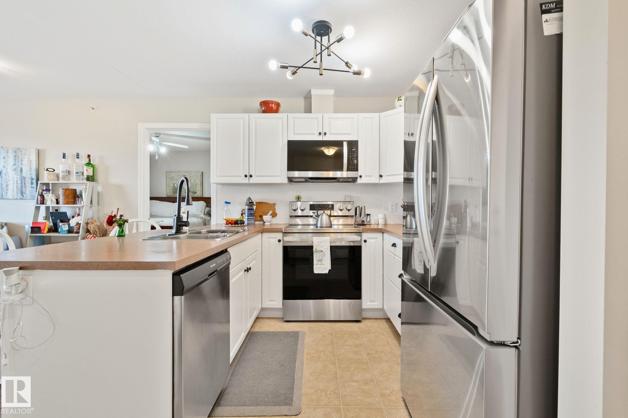 White cabinetry with black hardware, stainless steel appliances, a center island with sink, and a contemporary light fixture - #407 4309 33 St, Stony Plain, AB - Indoor Photo Showing Kitchen