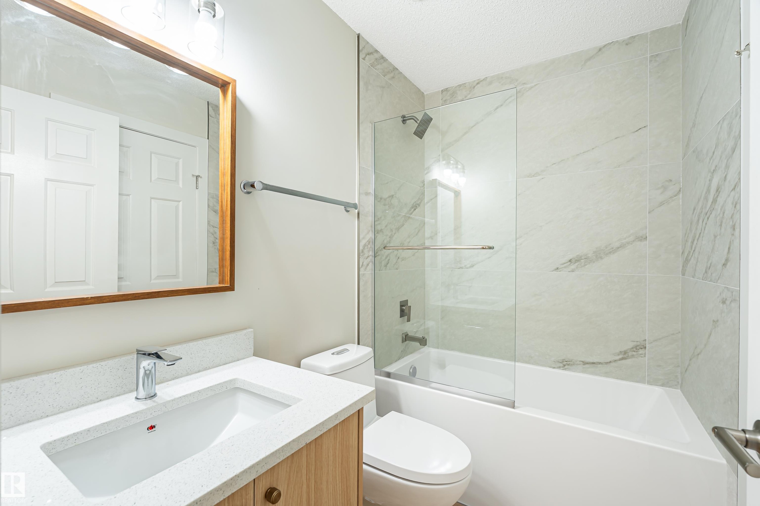 Bathroom featuring a vanity with an integrated sink and light wood-finish cabinetry, a framed mirror, a tub with a glass panel enclosure, and large format wall tile - #21 9375 172 St Nw, Edmonton, AB - Indoor Photo Showing Bathroom