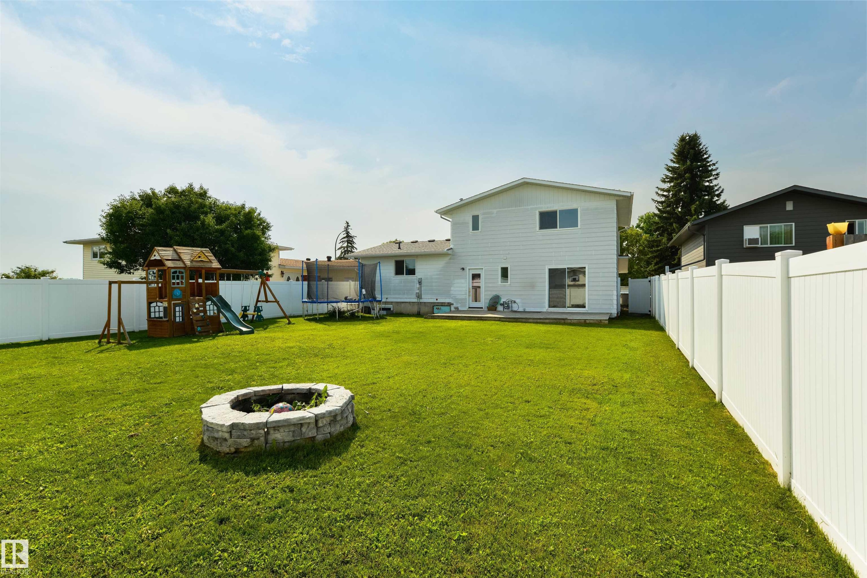 Expansive green lawn featuring a built-in stone fire pit, a white privacy fence, and a rear exterior with a sliding glass door - 4614 53 Ave, Calmar, AB - Outdoor With Backyard