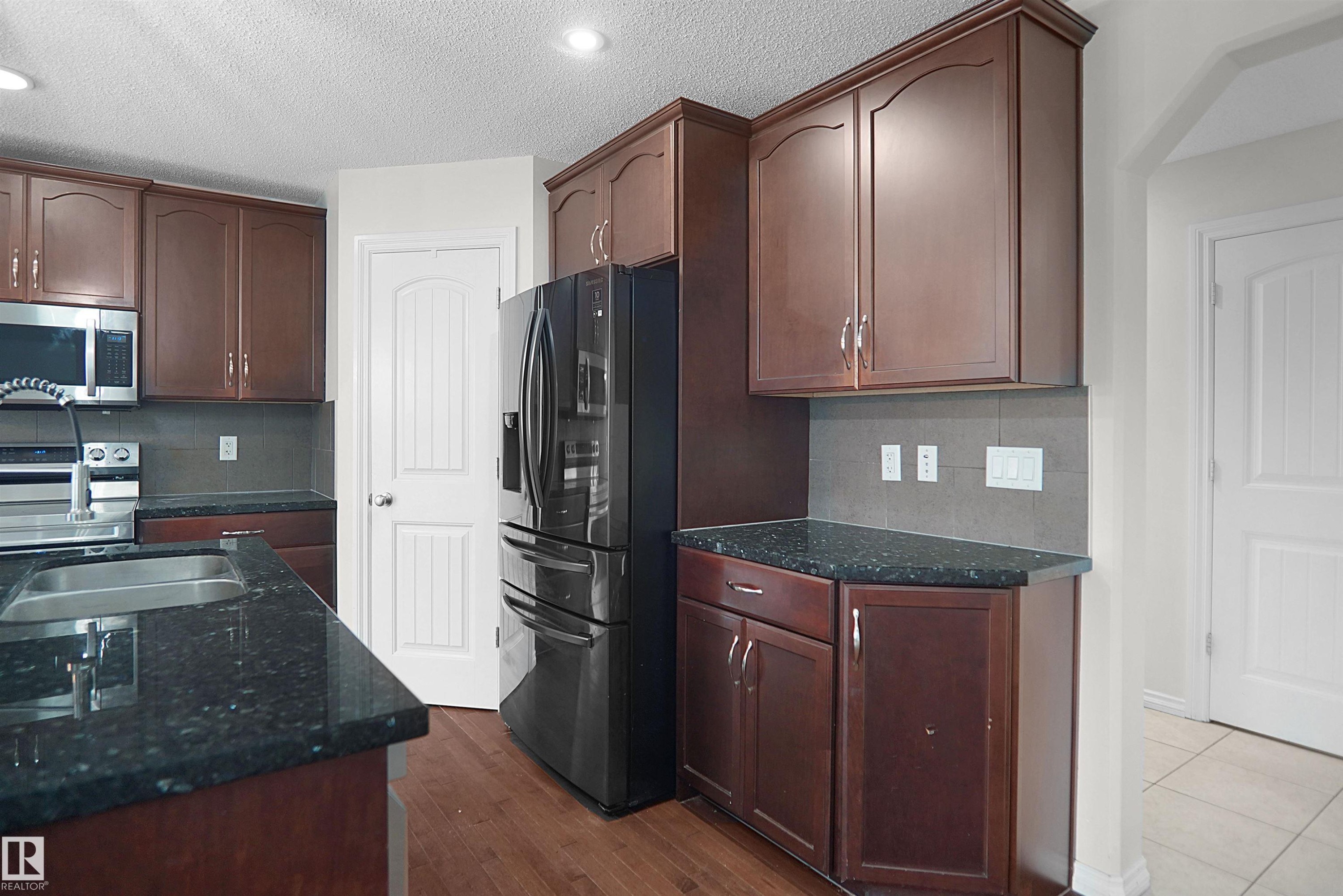 Dark wood-finish cabinetry with silver-tone hardware, dark countertop surfaces, stainless steel appliances, and wood-finish flooring - 8432 18 Avenue Sw, Edmonton, AB - Indoor Photo Showing Kitchen With Double Sink