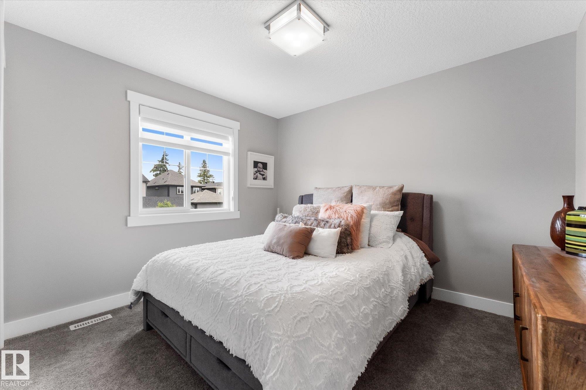 Bright secondary bedroom featuring a large double-hung window with natural light, soft gray walls, dark wood-finish flooring, and wood dresser with decorative accessories - 5616 Juchli Avenue Nw, Edmonton, AB - Indoor Photo Showing Bedroom