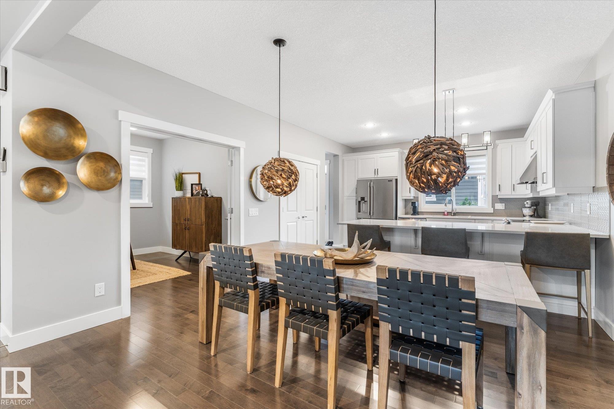 Open-concept dining and kitchen area featuring a wood-finish dining table with woven black leather dining chairs, pendant lighting with woven natural fiber shades, stainless steel kitchen appliances, white cabinetry, and hardwood flooring throughout - 5616 Juchli Avenue Nw, Edmonton, AB - Indoor Photo Showing Dining Room
