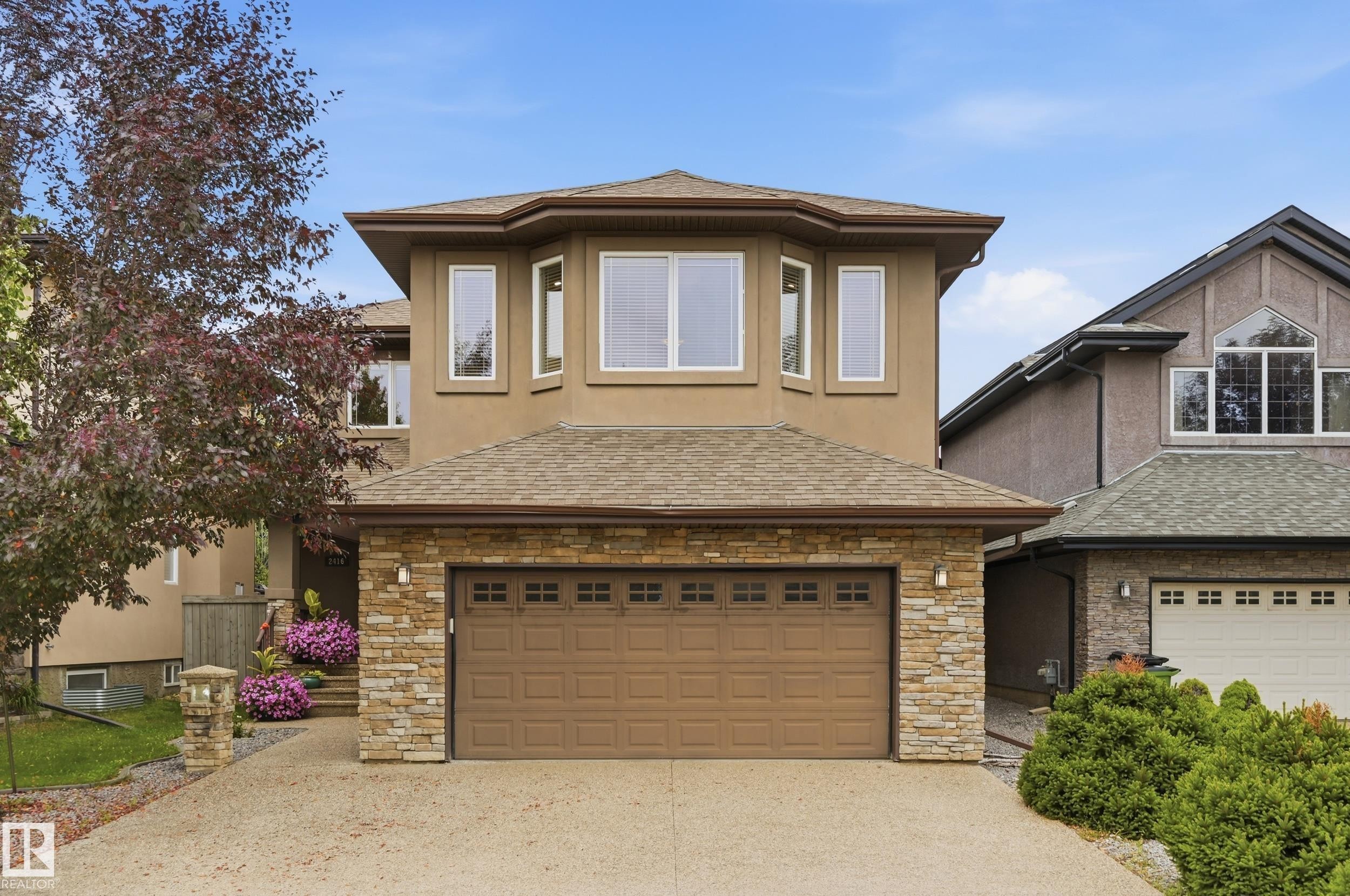Two-story home featuring a stone and stucco exterior with a covered entry, large picture windows, and a two-car garage door with paneled detailing - 2416 Warry Place Sw, Edmonton, AB - Outdoor