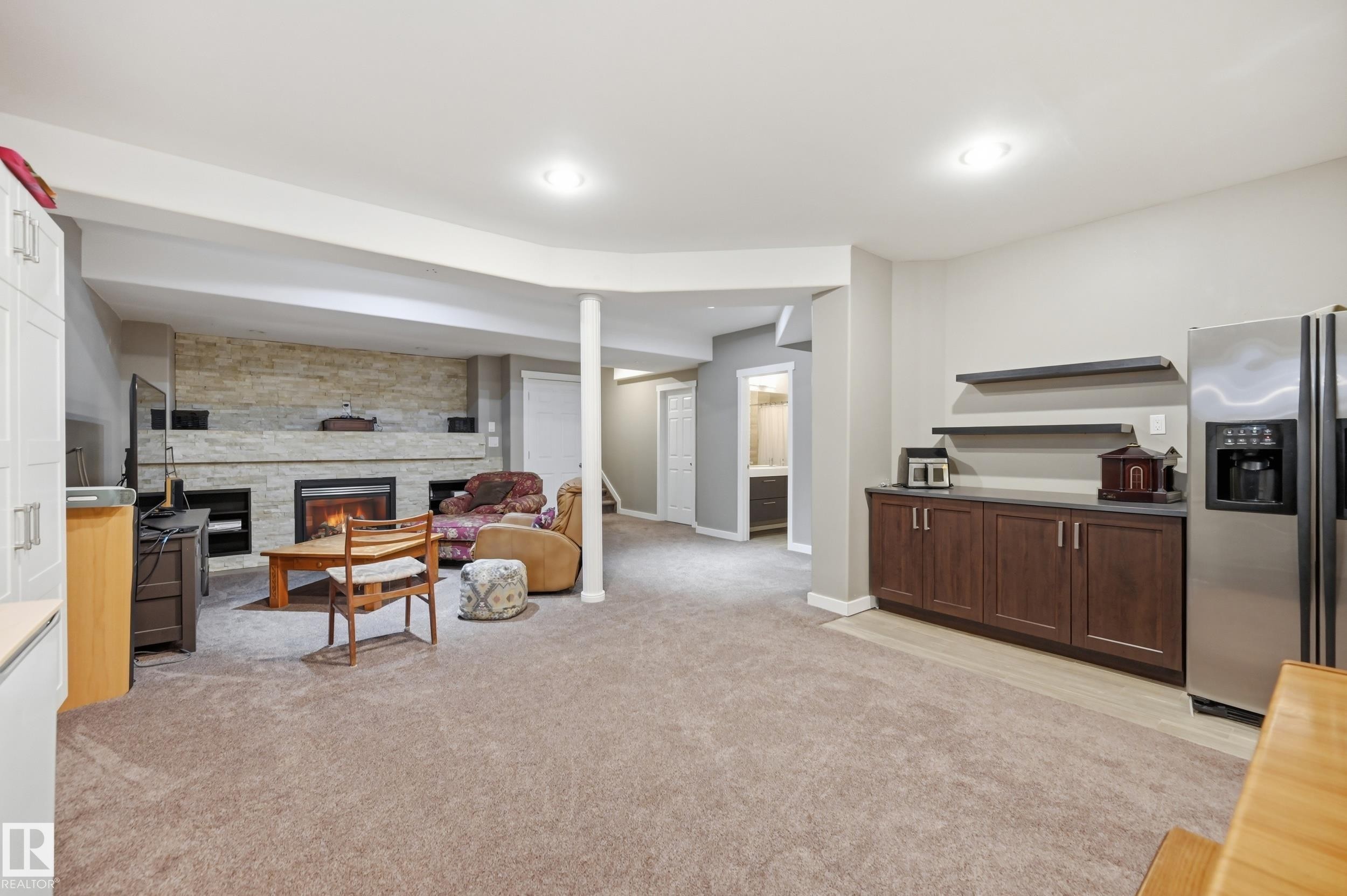 Spacious entertainment area featuring a stone-clad fireplace, carpeted flooring, built-in shelving, and a wet bar with dark wood cabinetry - 2416 Warry Place Sw, Edmonton, AB - Indoor