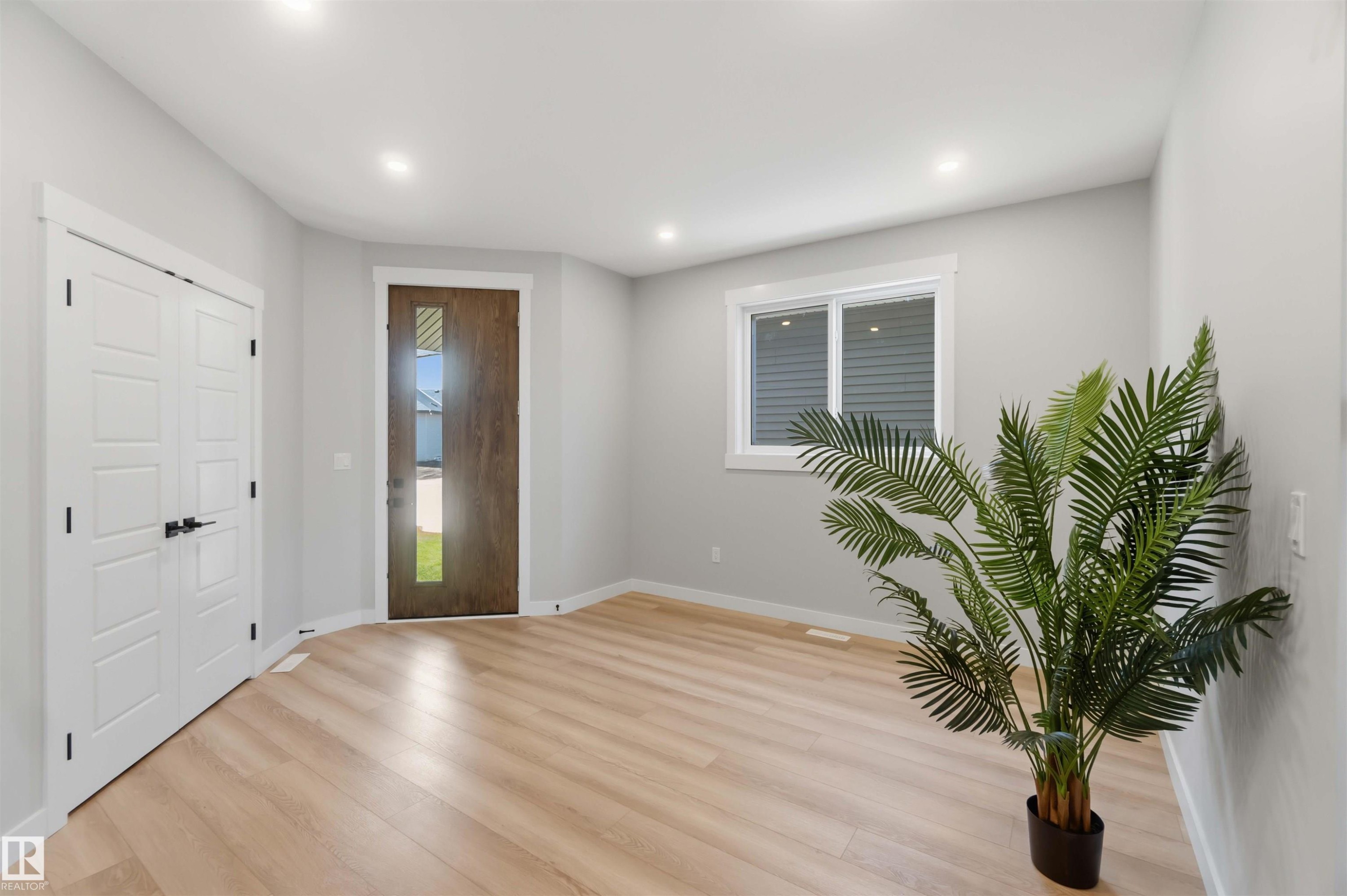 Wood-finish flooring, recessed ceiling lighting, a dark wood entry door with a vertical glass insert, and white bi-fold doors with black hardware - 28 Harley Way, Spruce Grove, AB - Indoor Photo Showing Other Room