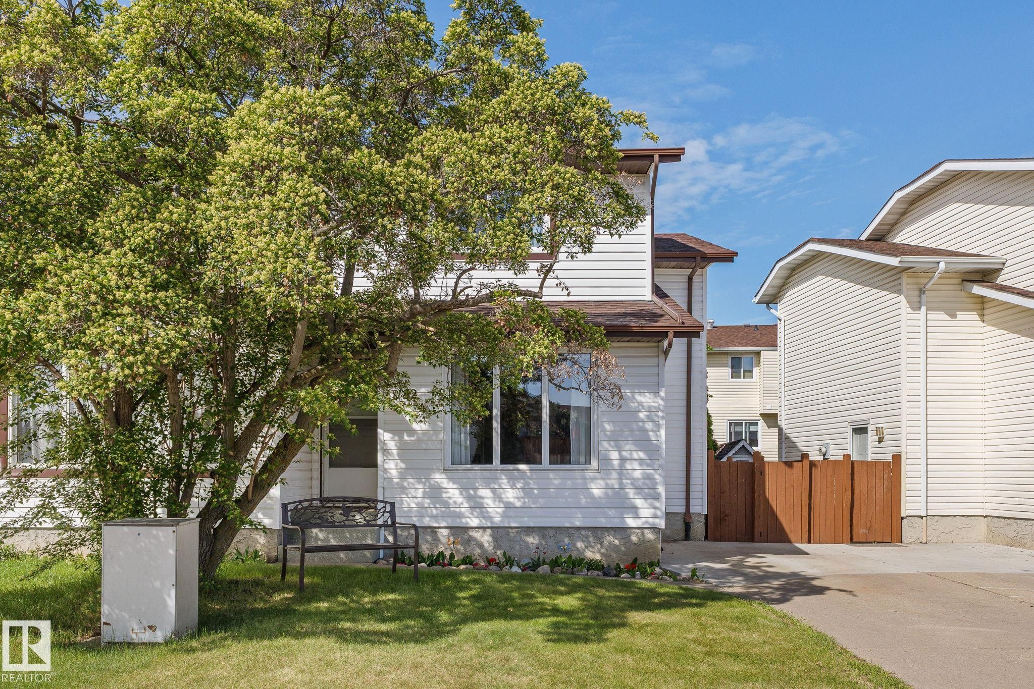 White siding exterior with a brown shingle roof, established front tree, and a concrete driveway leading to a privacy fence - 17132 103 Street Nw, Edmonton, AB