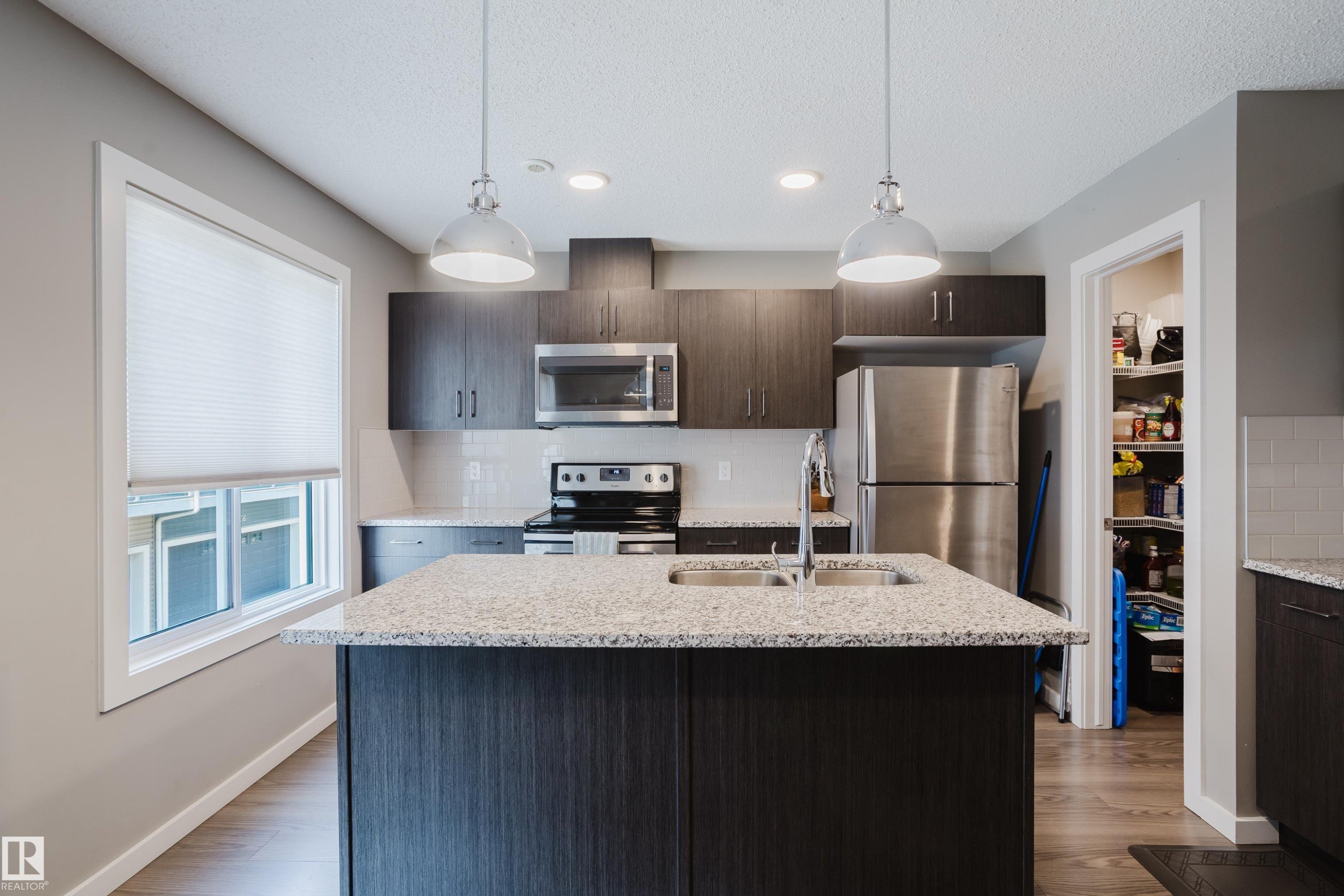 Modern kitchen featuring a central island with dual pendant lighting, stainless steel appliances, dark wood-finish cabinetry, light speckled countertops, and a full pantry - #28 2215 24 St Nw, Edmonton, AB - Indoor Photo Showing Kitchen With Double Sink With Upgraded Kitchen