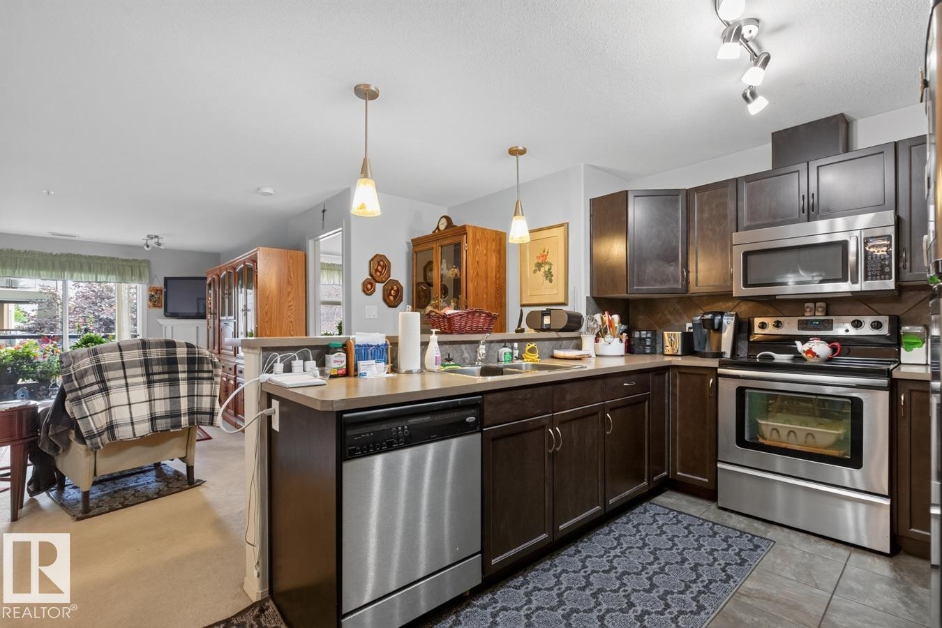 Kitchen featuring dark wood-finish cabinetry, stainless steel appliances, pendant lighting, and a neutral countertop - #210 7909 71 St Nw, Edmonton, AB - Indoor Photo Showing Kitchen With Double Sink