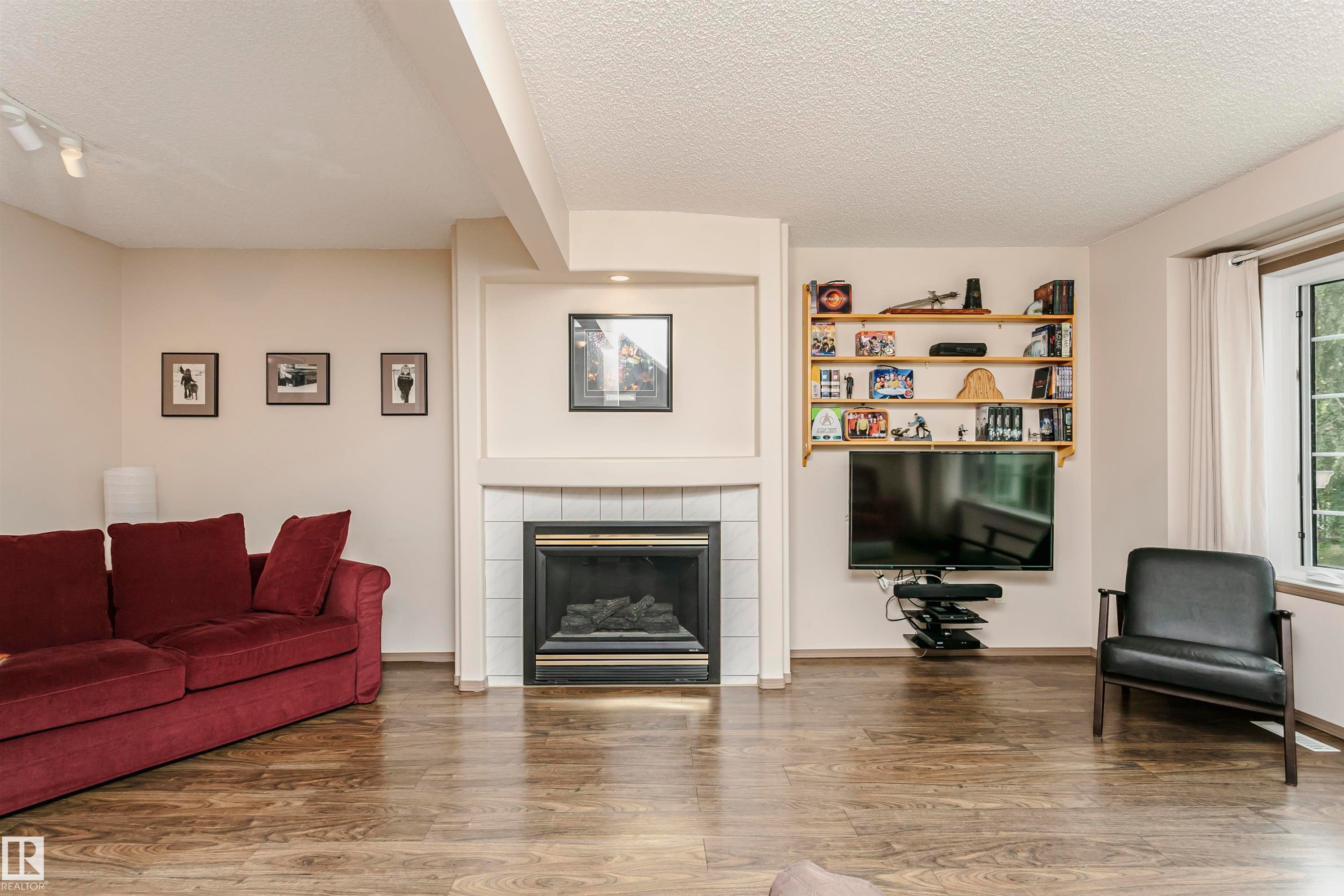Wood-finish flooring throughout, featuring a tiled fireplace with mantel, built-in shelving, and a prominent window - #42 9630 176 St Nw, Edmonton, AB - Indoor Photo Showing Living Room With Fireplace