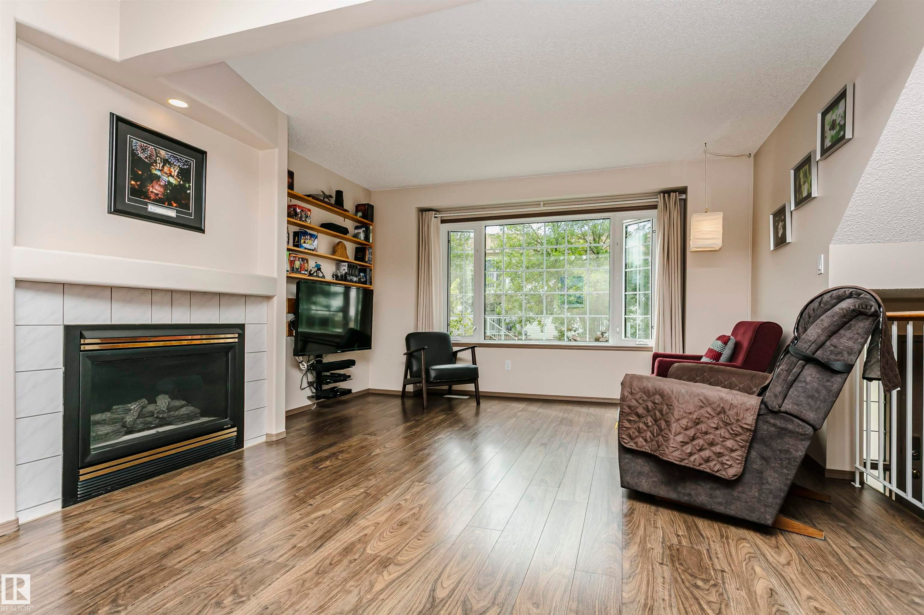 Living area featuring wood-finish flooring, a tiled fireplace with mantel, built-in shelving, a large bay window, and a white railing - #42 9630 176 St Nw, Edmonton, AB - Indoor Photo Showing Living Room With Fireplace
