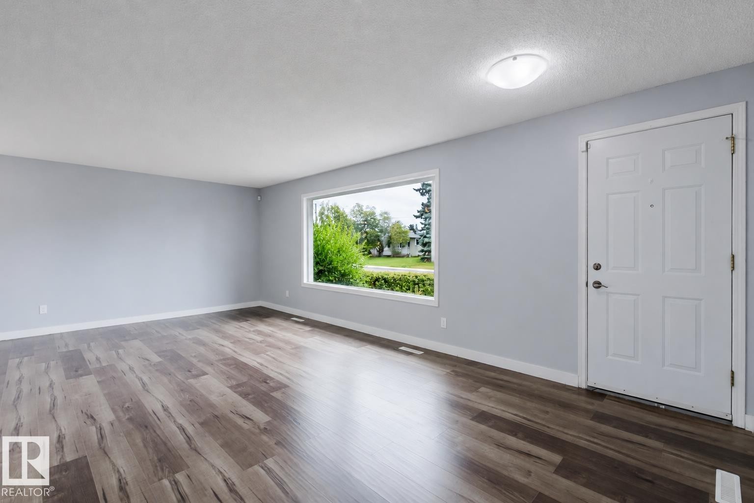 Expansive room featuring wood-finish flooring, a large window, a white entry door, and a flush-mount ceiling light - 11420 46 Avenue Nw, Edmonton, AB - Indoor