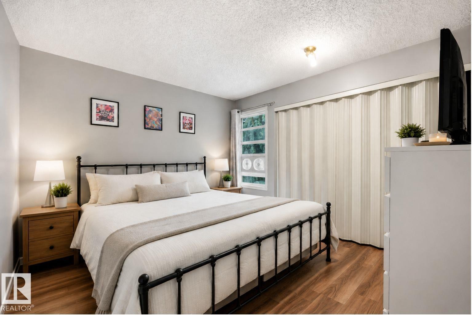Master bedroom featuring a black metal bed frame, wood-finish flooring, soft neutral bedding, dual nightstands with integrated lighting, framed artwork, vertical sheer window treatments, and a walk-in closet with built-in shelving and storage organization - #44 1415 62 St Nw, Edmonton, AB - Indoor Photo Showing Bedroom