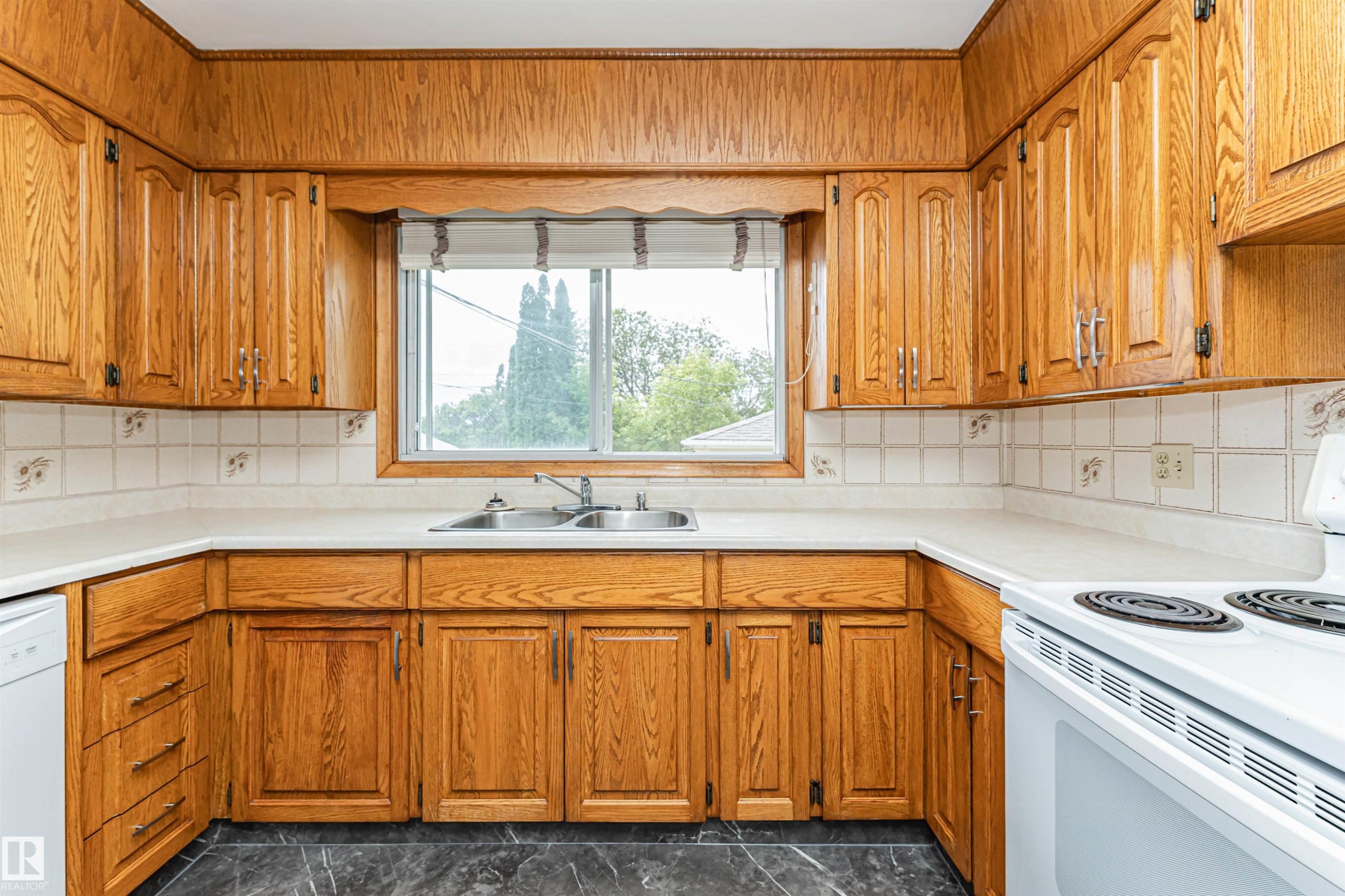 U-shaped kitchen with extensive wood cabinetry, light-toned countertops, a double basin stainless steel sink, tile backsplash, and dark-toned flooring - 11139 62 Avenue Nw, Edmonton, AB - Indoor Photo Showing Kitchen With Double Sink