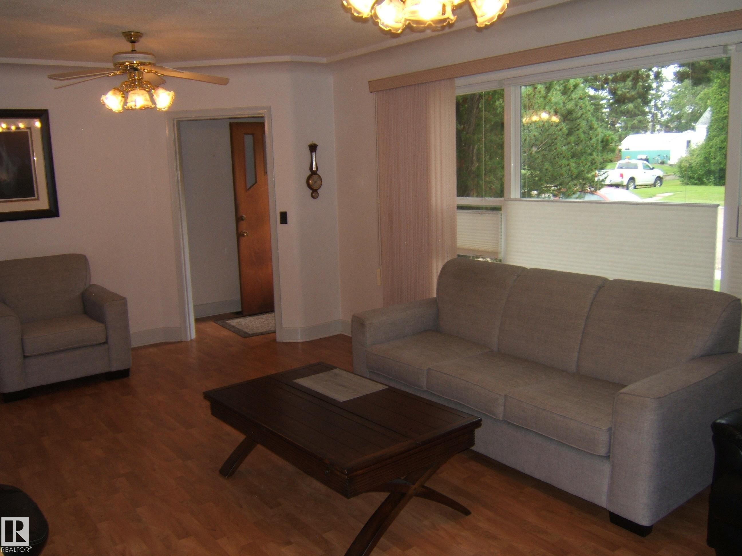 Bright living room showcasing wood-finish flooring, a three-cushion upholstered sofa, accent armchair, and dark wood coffee table - 4809 47A Street, Two Hills, AB - Indoor Photo Showing Living Room