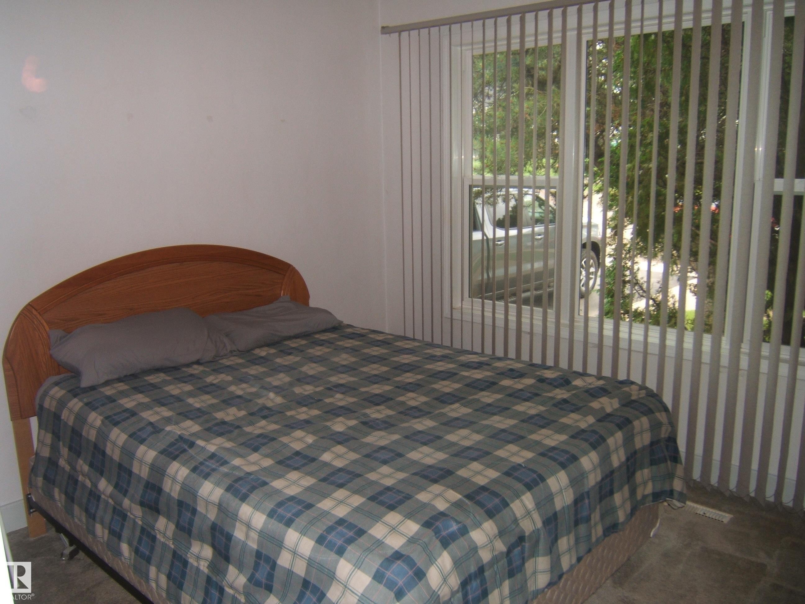 Comfortable bedroom featuring a wood-finish bed frame, plaid bedding, and vertical blinds - 4809 47A Street, Two Hills, AB - Indoor Photo Showing Bedroom