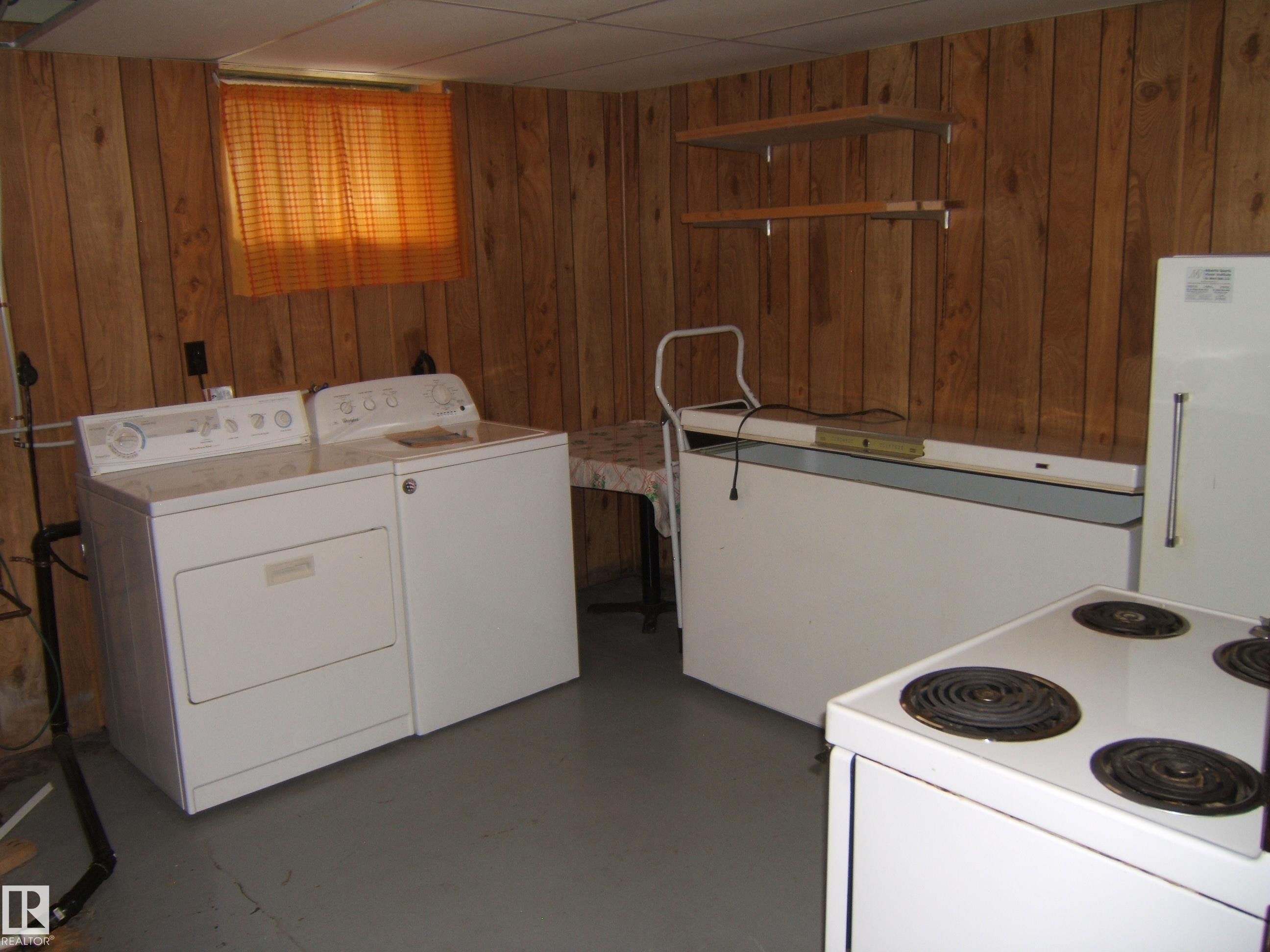 Utility room featuring wood-paneled walls, stacked washer and dryer unit, electric range with coil burners, and a standalone bathtub with handrail - 4809 47A Street, Two Hills, AB - Indoor Photo Showing Laundry Room