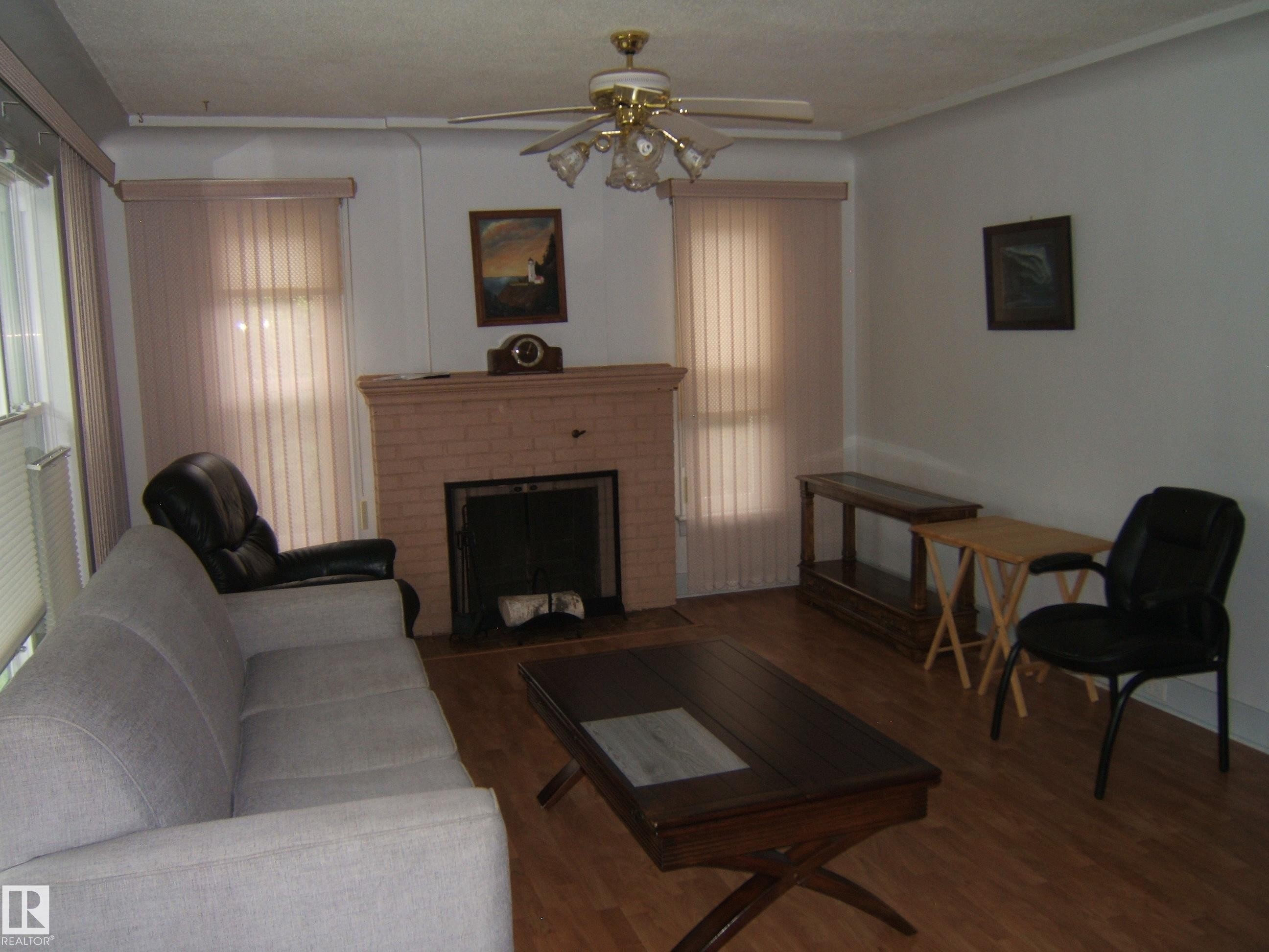 Living area featuring a brick fireplace, wood-finish flooring, ceiling fan with integrated lighting, and vertical window blinds - 4809 47A Street, Two Hills, AB - Indoor Photo Showing Living Room With Fireplace